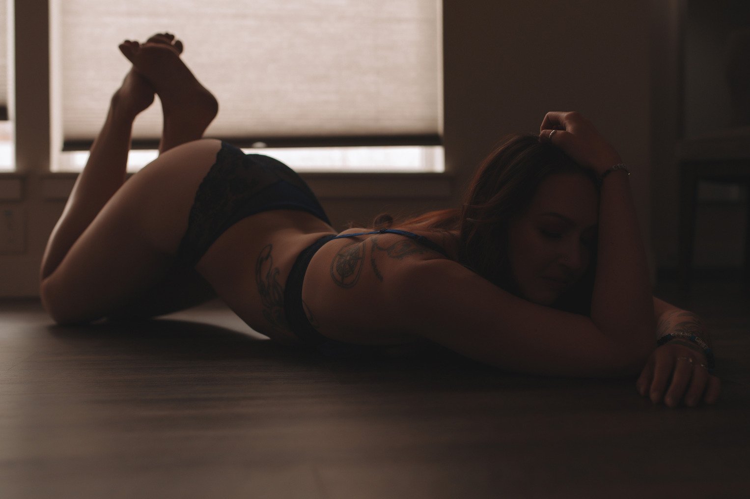 A woman in dark lingerie lies on her stomach on a wooden floor in a dimly lit room, capturing the intimate feel of boudoir photos, with one arm resting under her forehead and her legs bent upward.