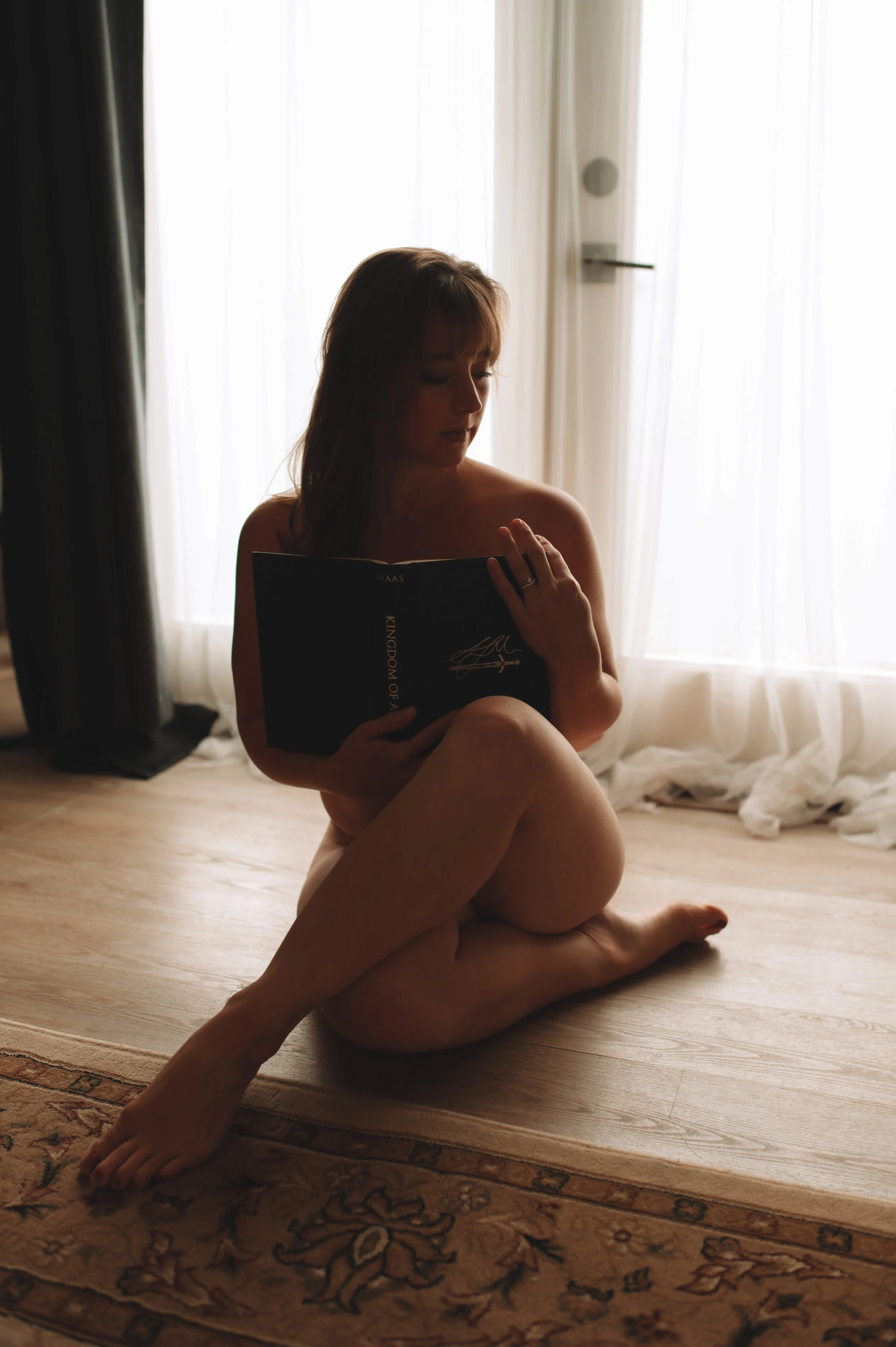 A woman sitting on the floor near a window, reading a book, with soft natural light illuminating her silhouette.