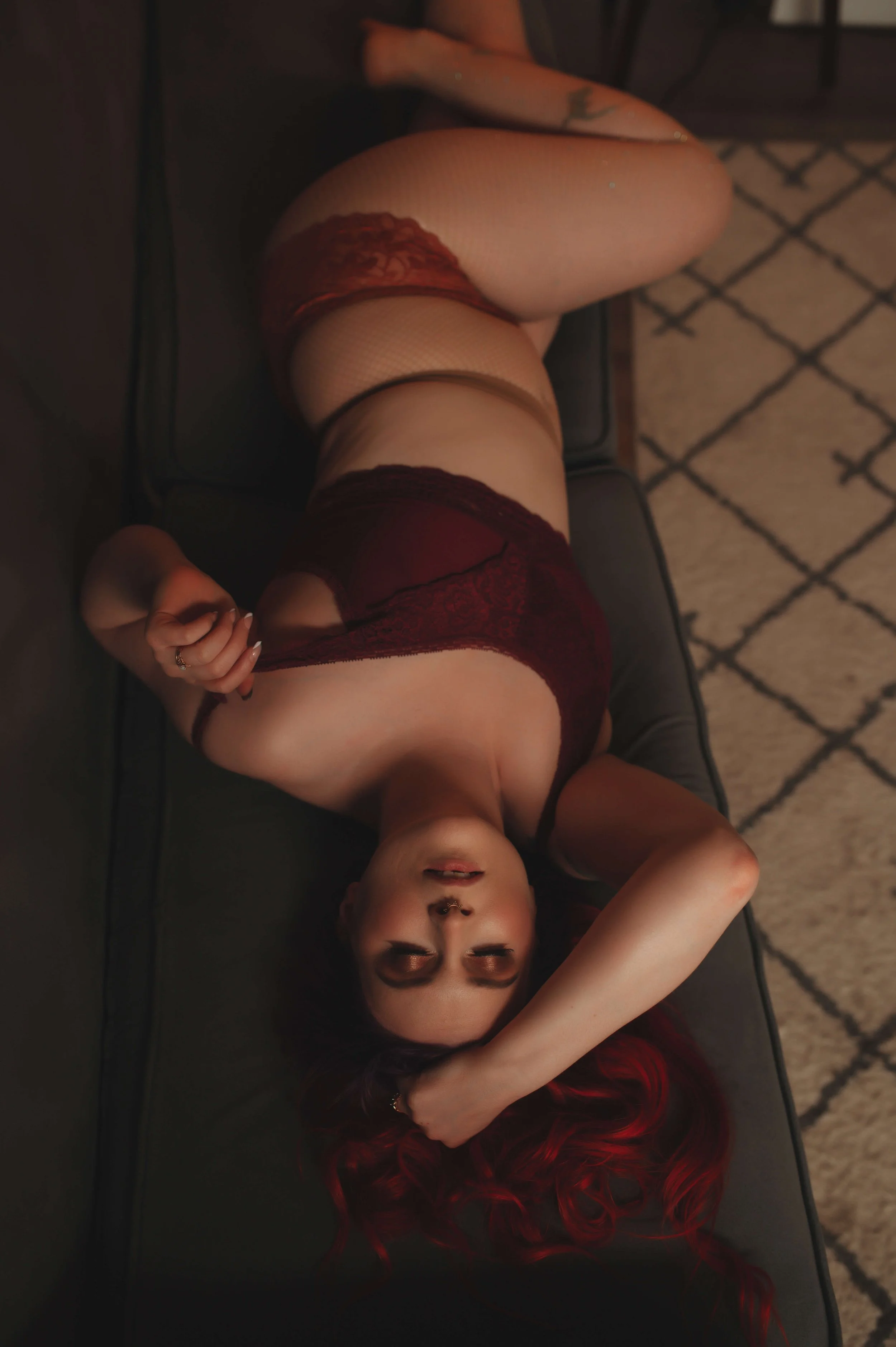 A woman with red hair lying on her back on a dark couch, wearing burgundy lace lingerie and fishnet stockings, with her eyes closed.