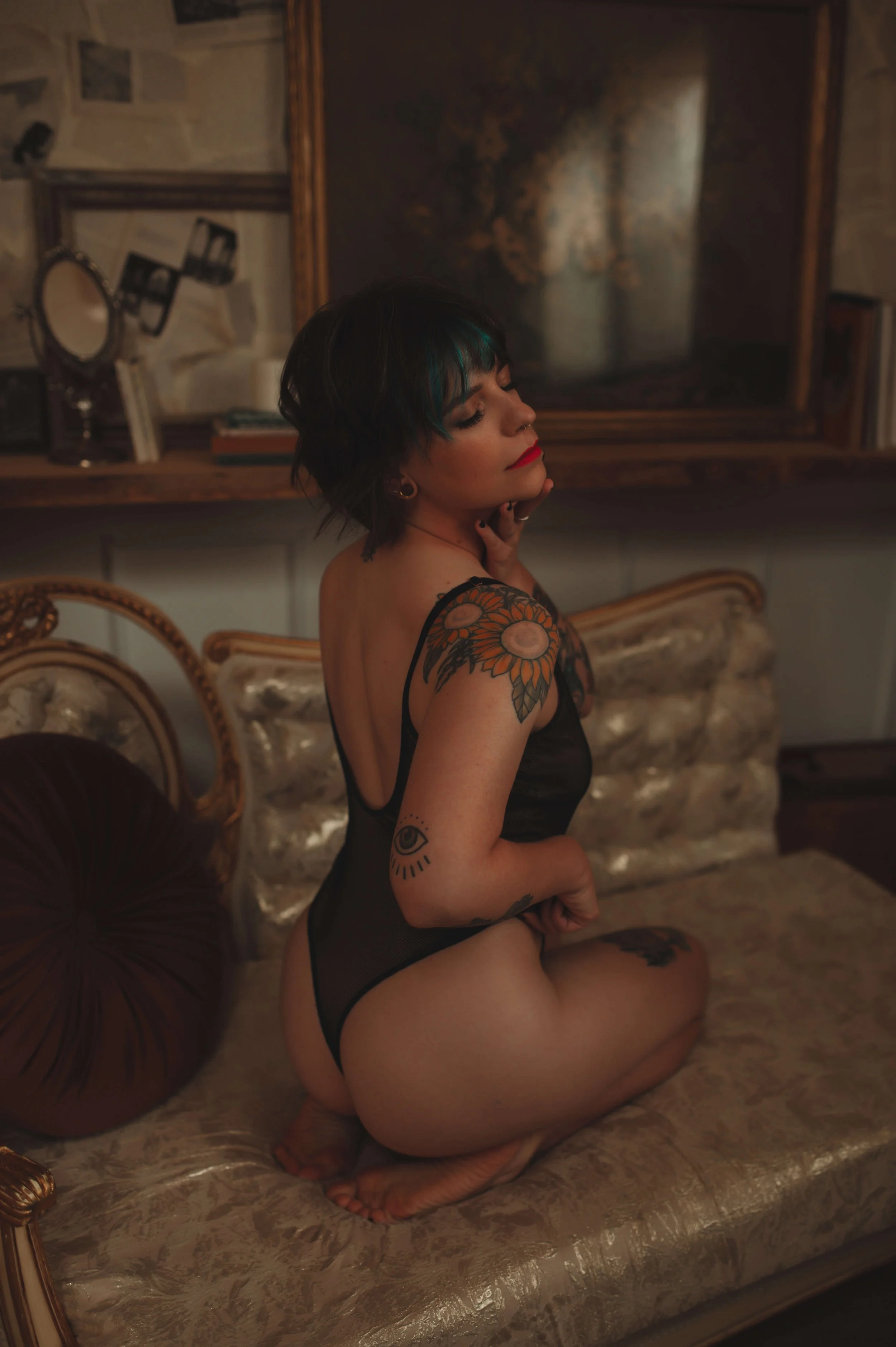 A woman with short dark hair and colorful tattoos on her arm and leg, sitting on her knees on a vintage sofa in a dimly lit room with dark, abstract art on the wall behind her.