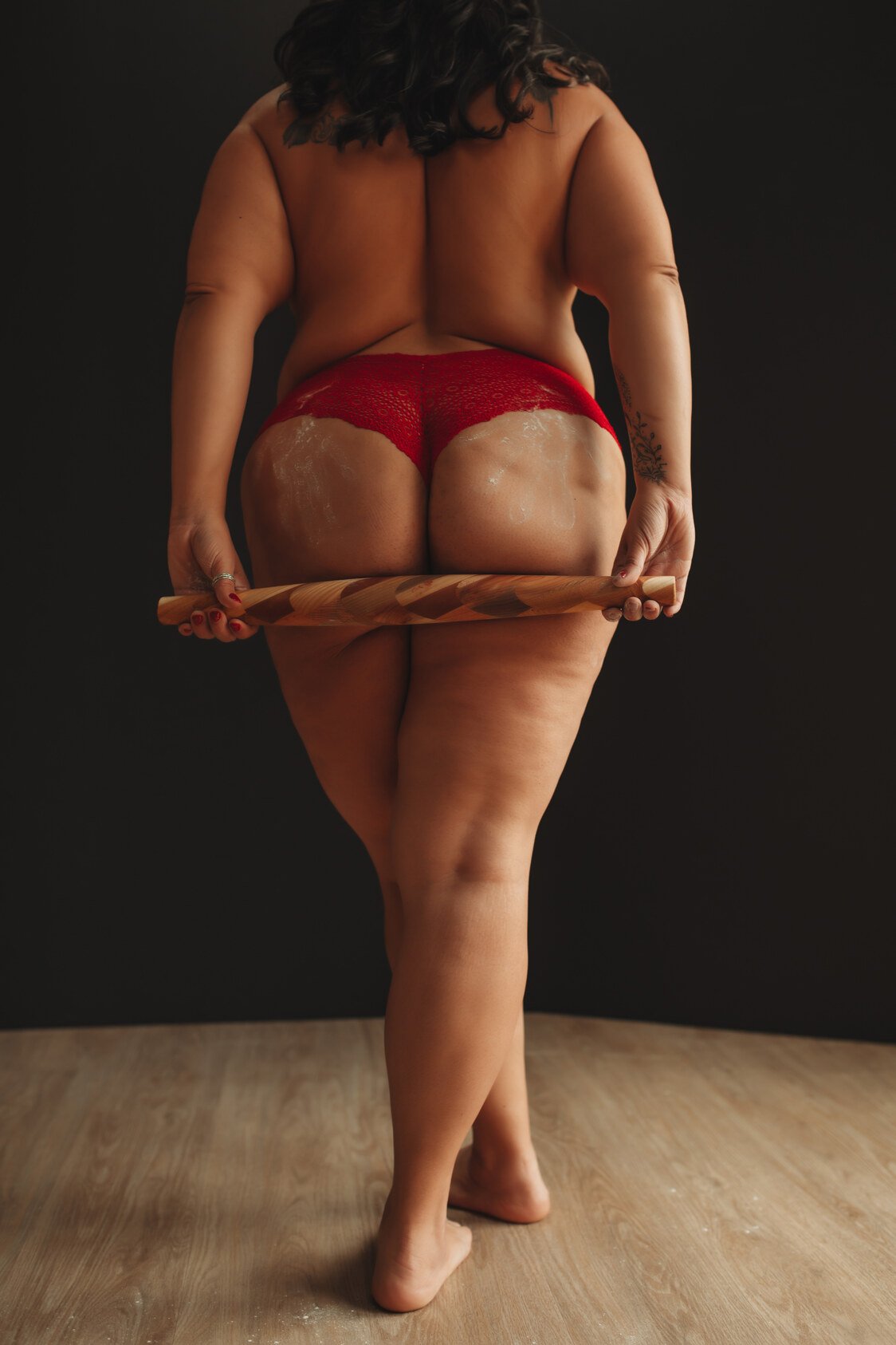 A person with long dark hair, wearing red lingerie and holding a wooden rolling pin behind their back, stands barefoot on a wooden floor against a dark background—capturing the intimate style of boudoir photos.