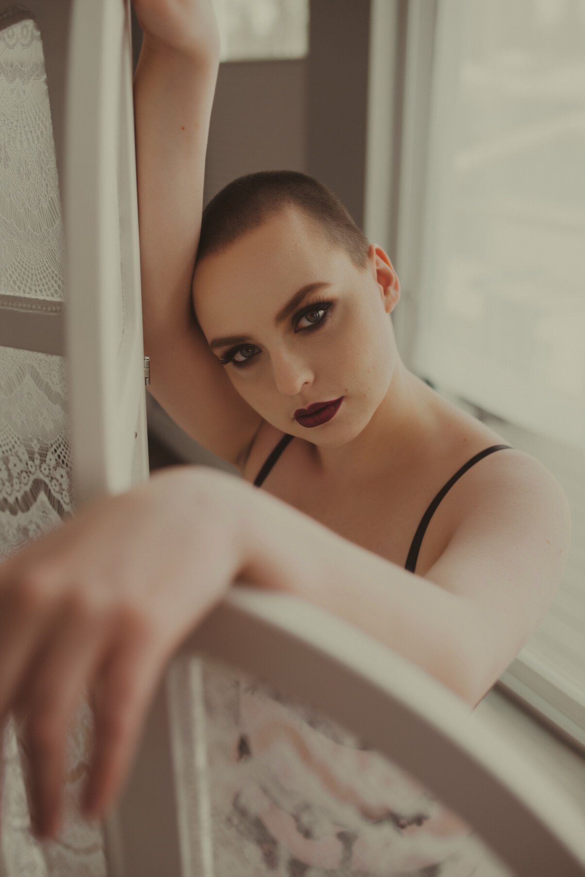 A person with a shaved head and dramatic makeup in lingerie leans against a lace-covered screen, gazing intensely at the camera. The scene, bathed in soft natural light, captures the intimate feel of boudoir photos with personalized attention to deta