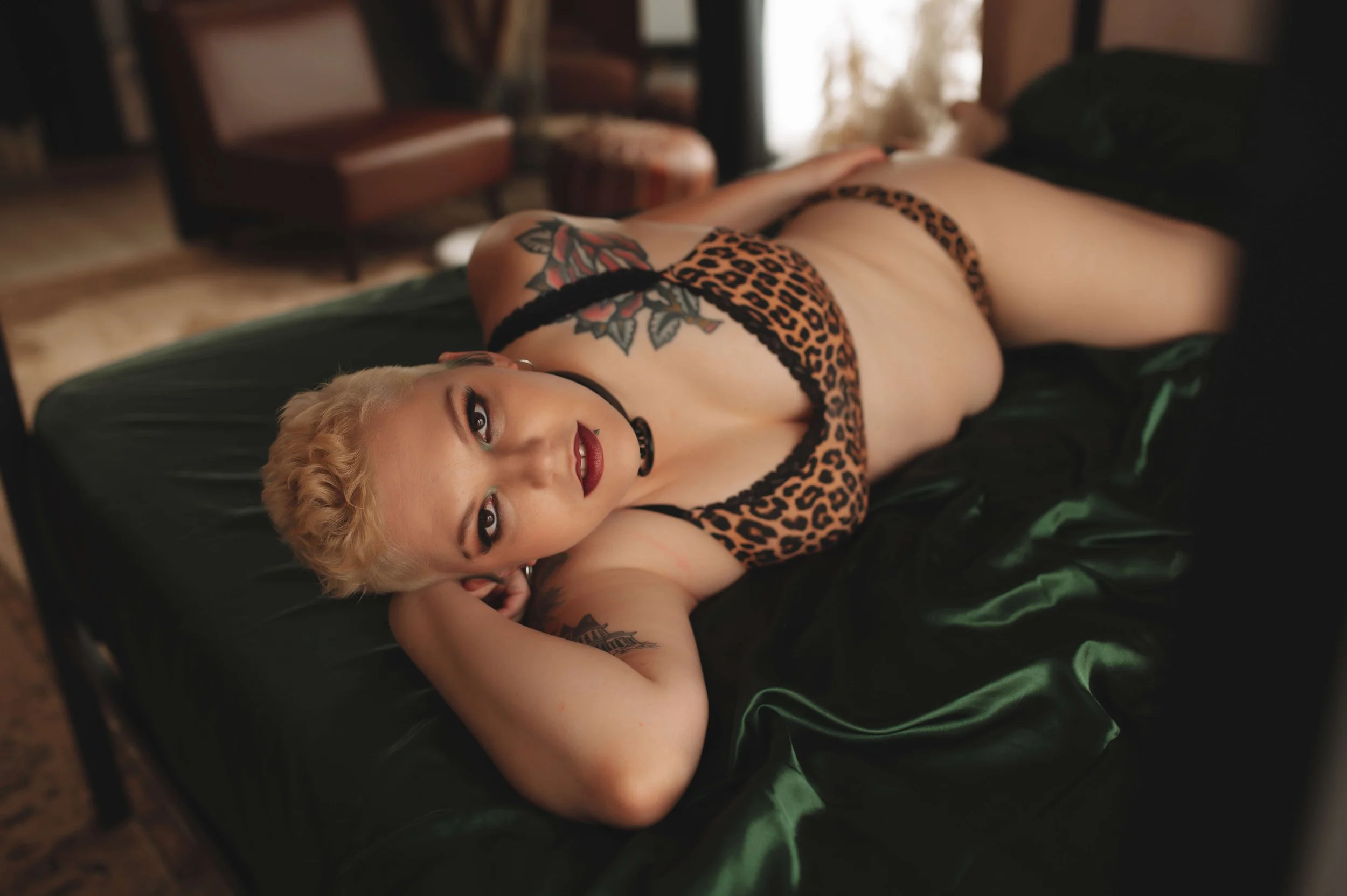 A woman with short blonde hair lying on a bed with dark green sheets, wearing leopard print lingerie and a choker. She has visible tattoos on her shoulder and arm, and is looking at the camera with a neutral expression.