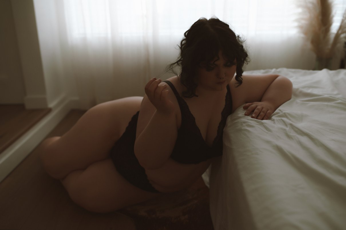 A woman with dark curly hair, wearing black lingerie, sits on the floor next to a bed with white sheets in a softly lit room with sheer curtains—capturing the intimate elegance of boudoir photos.