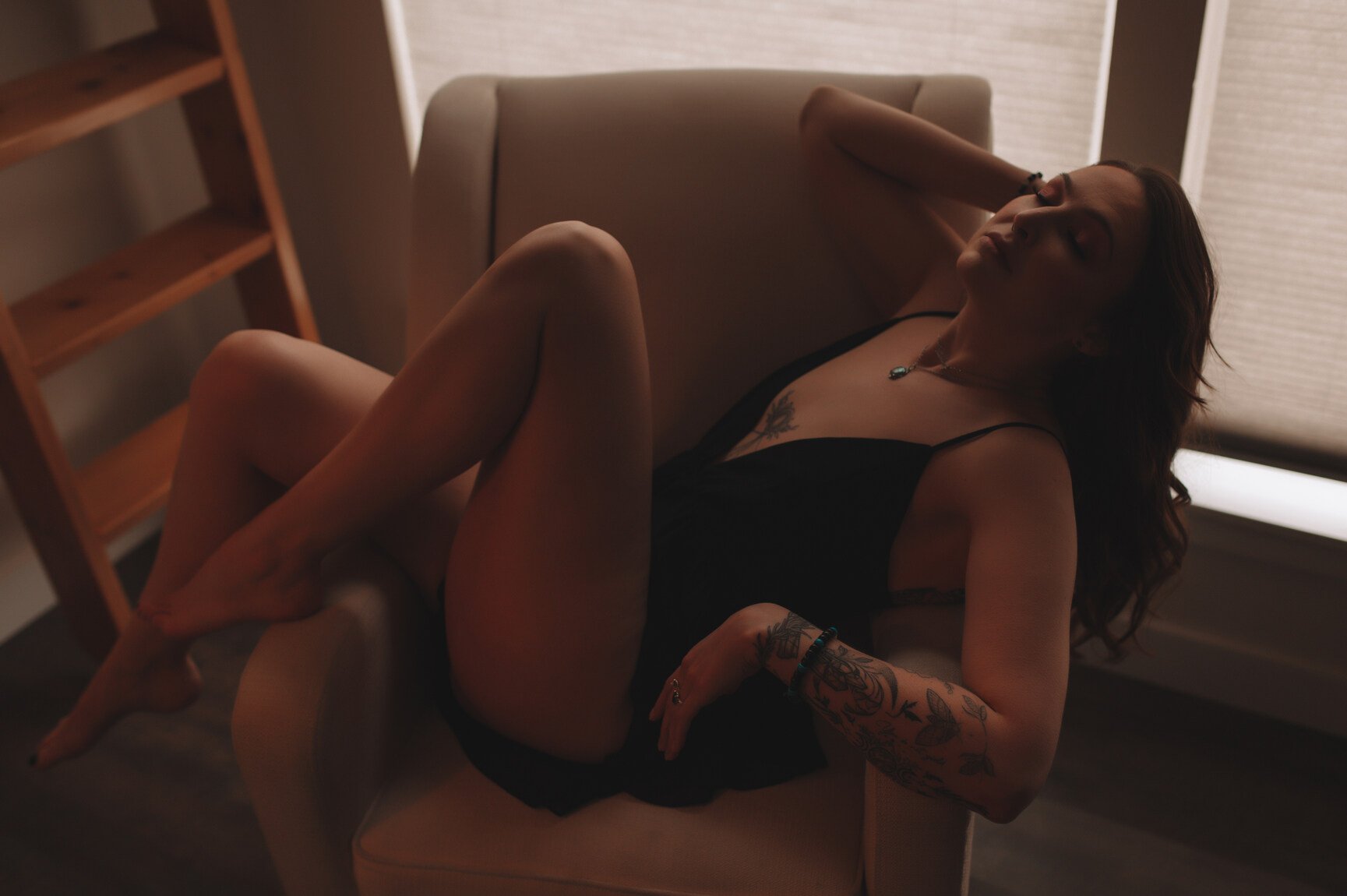 Woman with long brown hair and tattoos wears black romper and reclines over the arm of a cream colored arm chair
