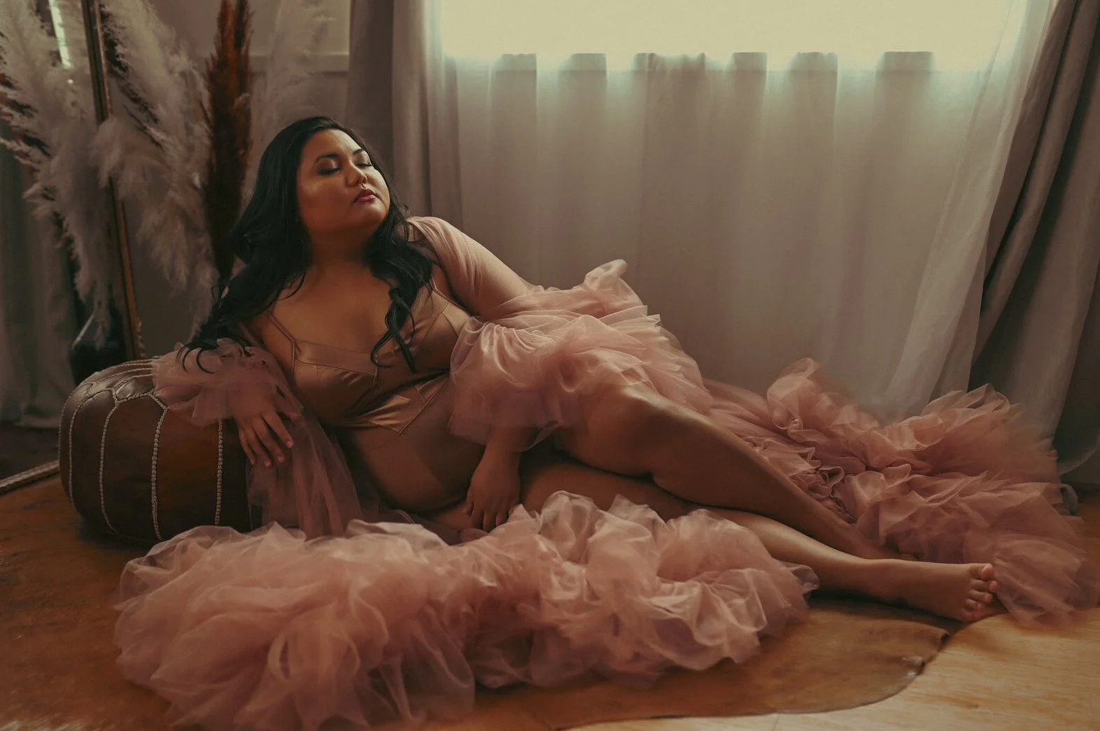 A woman with long dark hair reclining on a brown leather pouf, wearing a pink tulle dress, in a softly lit room with curtains and feathered decor in the background.