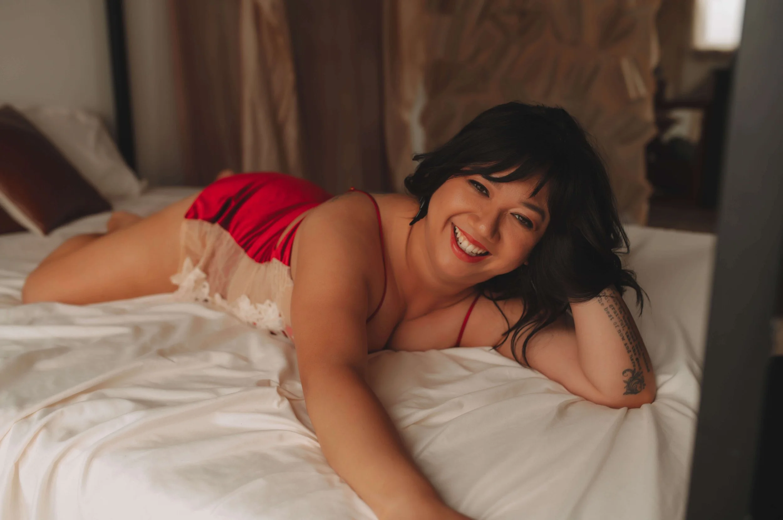 A woman with black hair and tattoos lying on her side on a bed, smiling and looking at a screen.