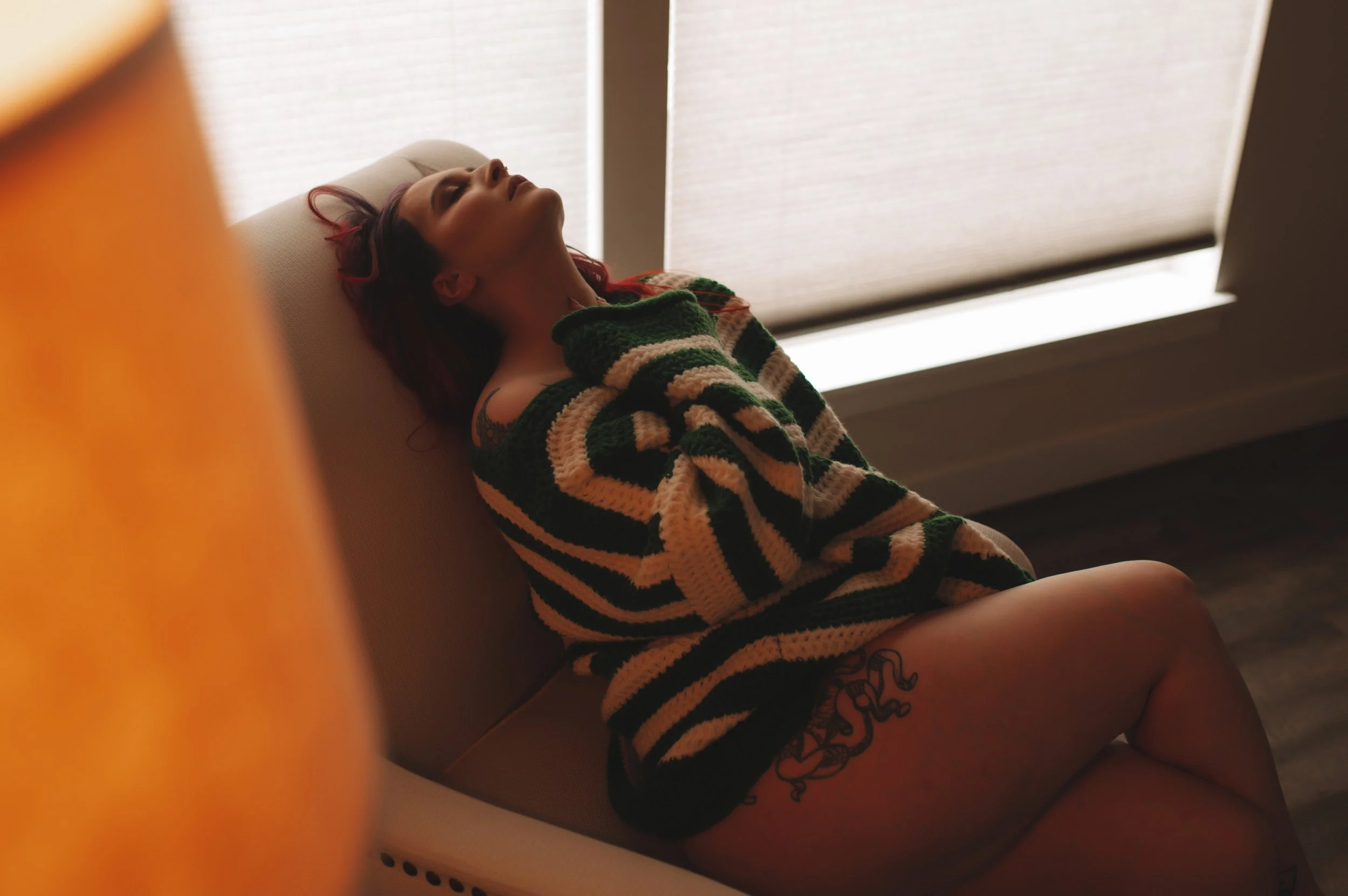 A woman with red hair lying on a beige lounge chair near a window with closed blinds, wearing an oversized striped sweater.