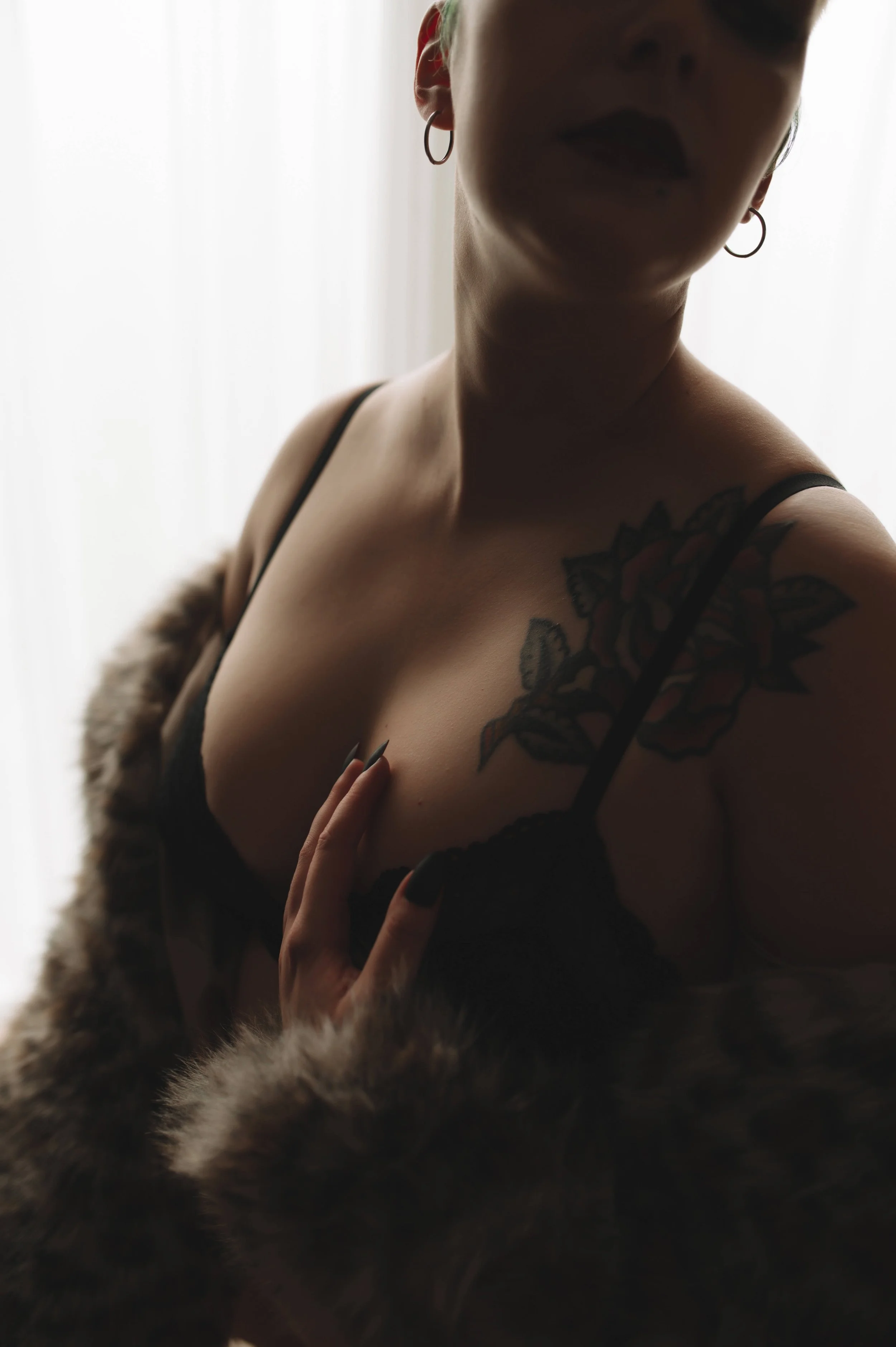A woman with tattoos and hoop earrings wearing a black lace top, standing against a light background, with a fur coat draped over her.