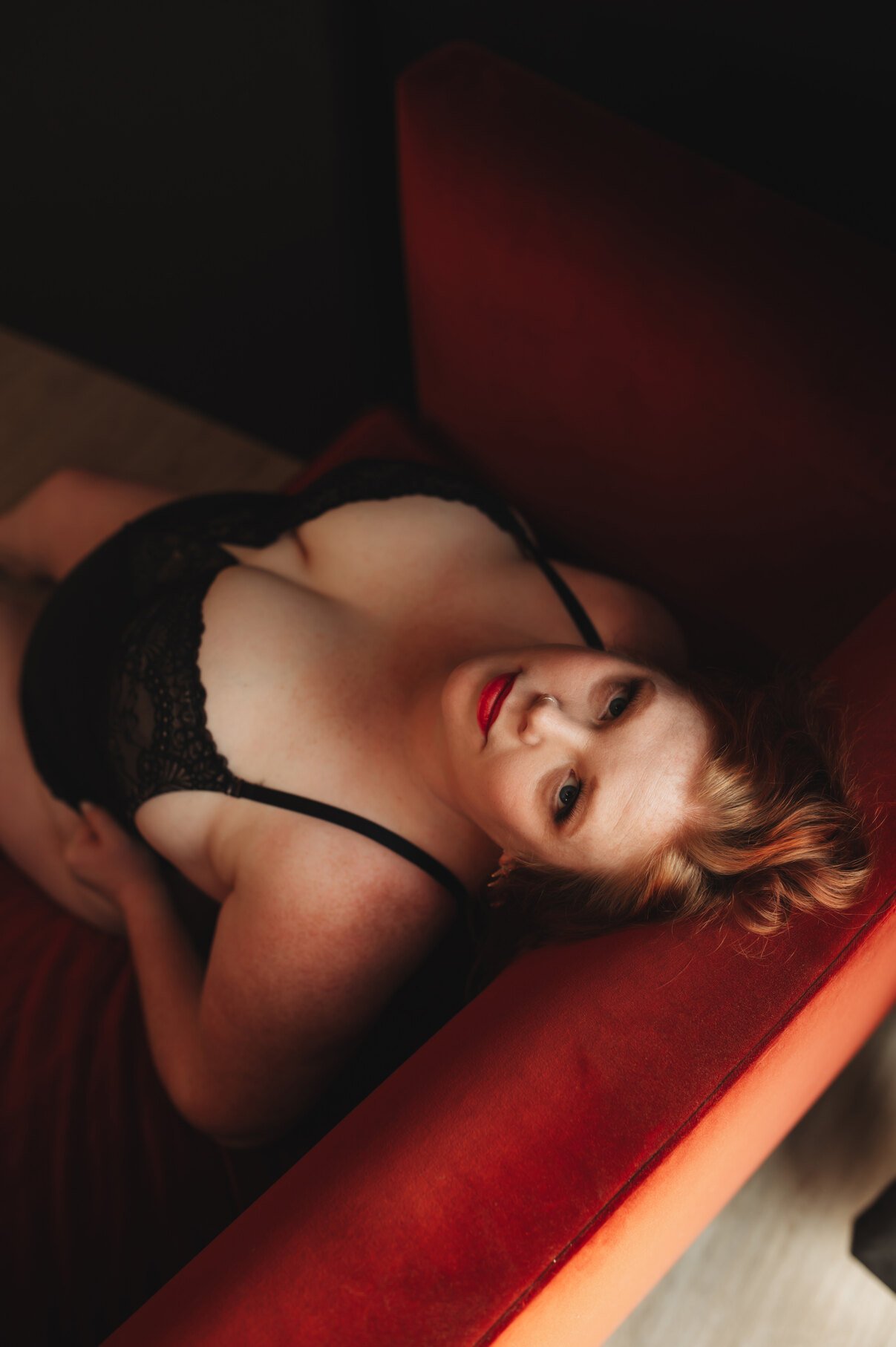 A woman with red hair and red lipstick reclines on a red chair, looking up at the camera. She wears black lace lingerie, while soft lighting highlights her face and shoulders for stunning boudoir photos.