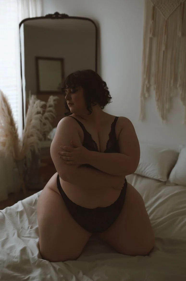 A woman in black lingerie kneeling on a bed with her arms crossed over her chest in a softly lit bedroom.