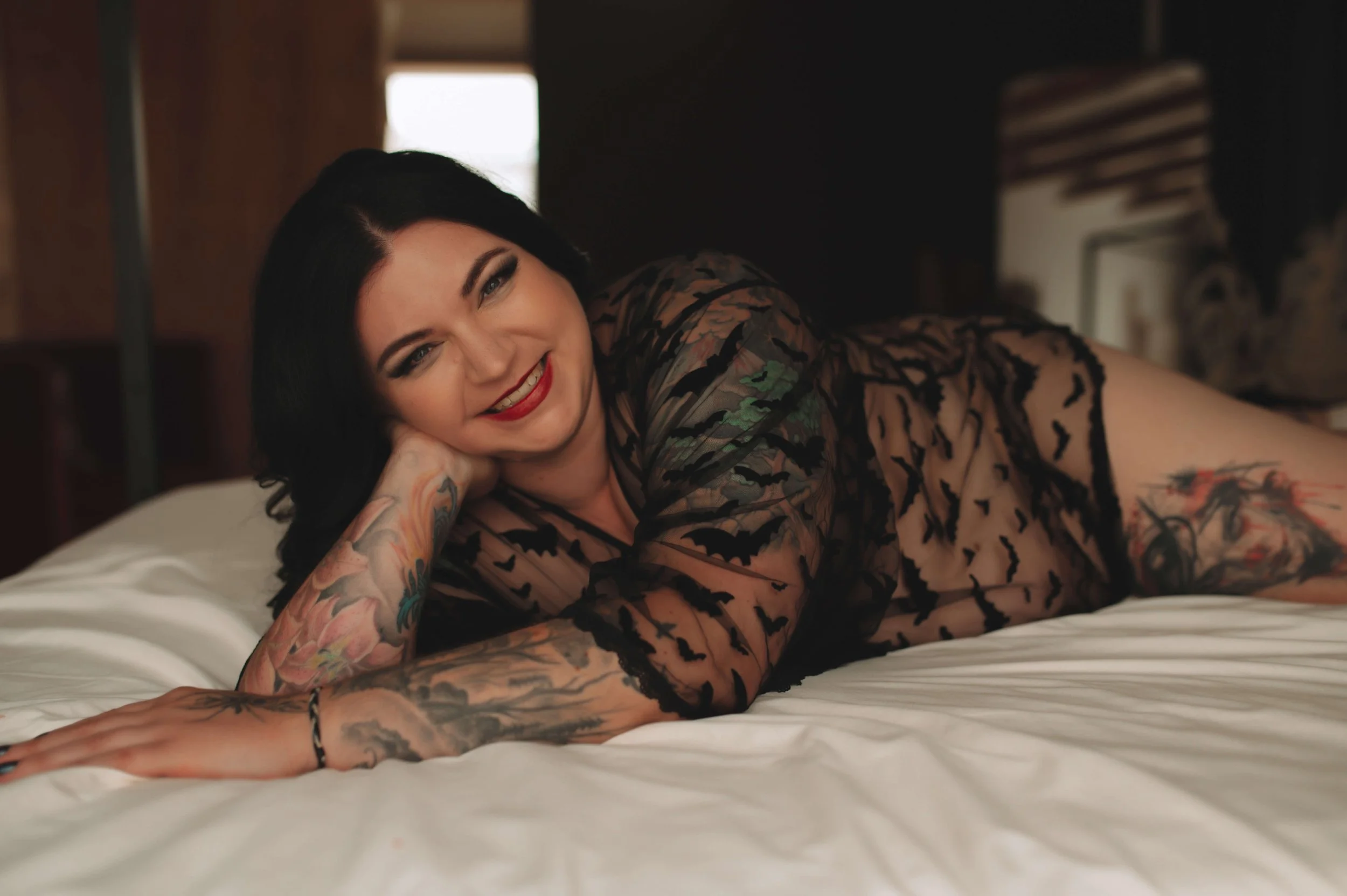 A woman with black hair, red lipstick, and tattoos, lying on a bed wearing a black sheer dress with bat patterns, smiling at the camera.