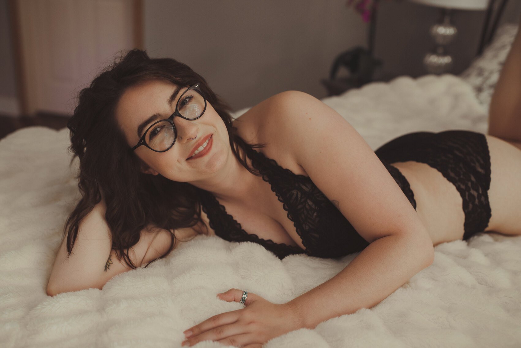 A woman with dark hair and glasses, wearing black lace lingerie, lies on her side on a fluffy white blanket, smiling at the camera in this intimate boudoir photo.