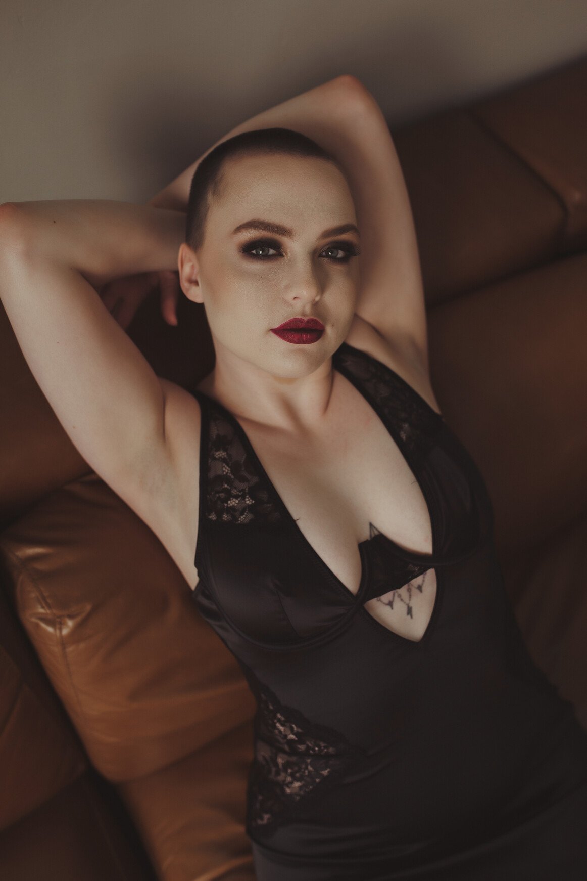 A person with a shaved head is reclining on a brown leather couch, wearing black lace-trimmed lingerie. They have bold makeup, with dark eyeshadow and red lipstick, capturing the allure of boudoir photos as their arms rest behind their head.