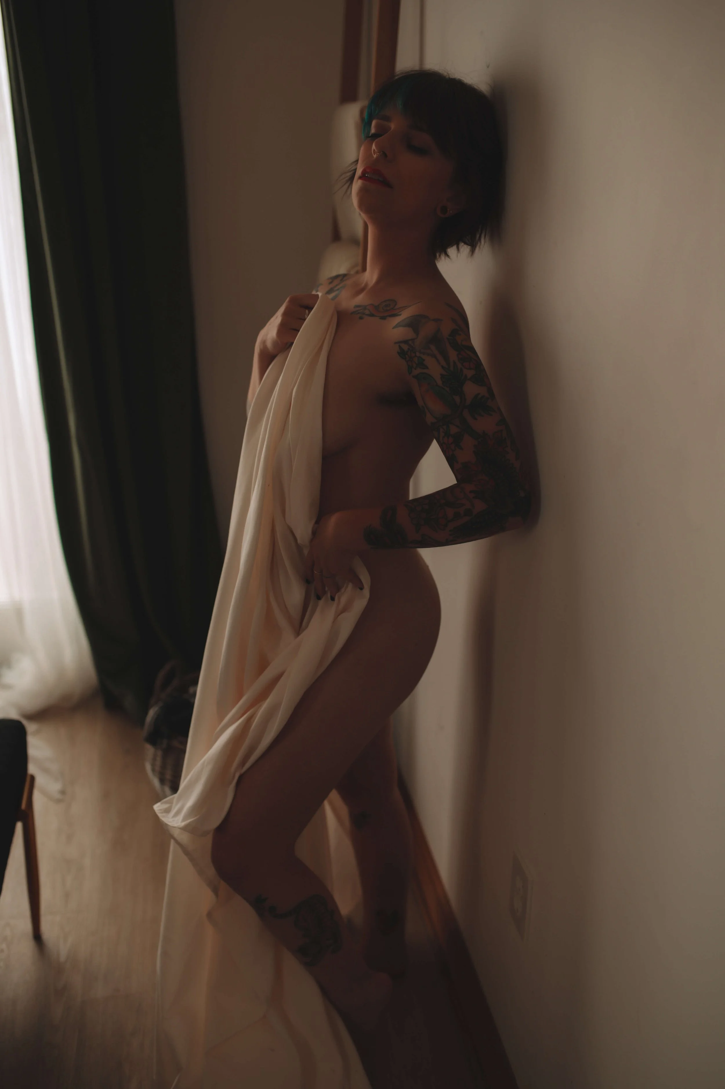 A woman with short dark hair and tattoos is posing nude against a wall in a dimly lit room, holding a white sheet partially covering her body.