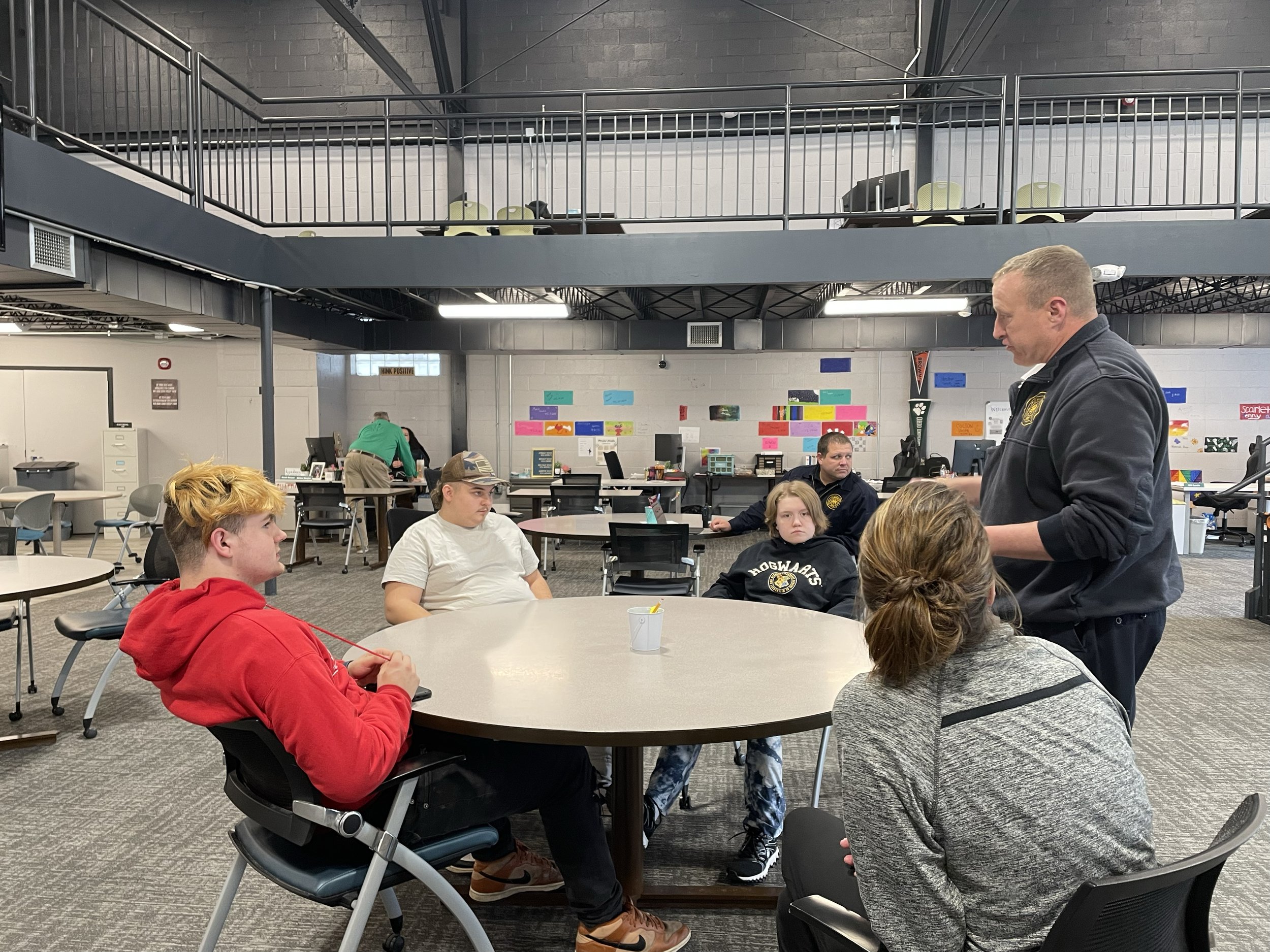 A group of people sitting at round tables in a modern classroom or meeting space, engaged in a discussion or lesson. Some are seated while one person is standing and talking, wearing casual attire. The room has an open layout with a mezzanine level a