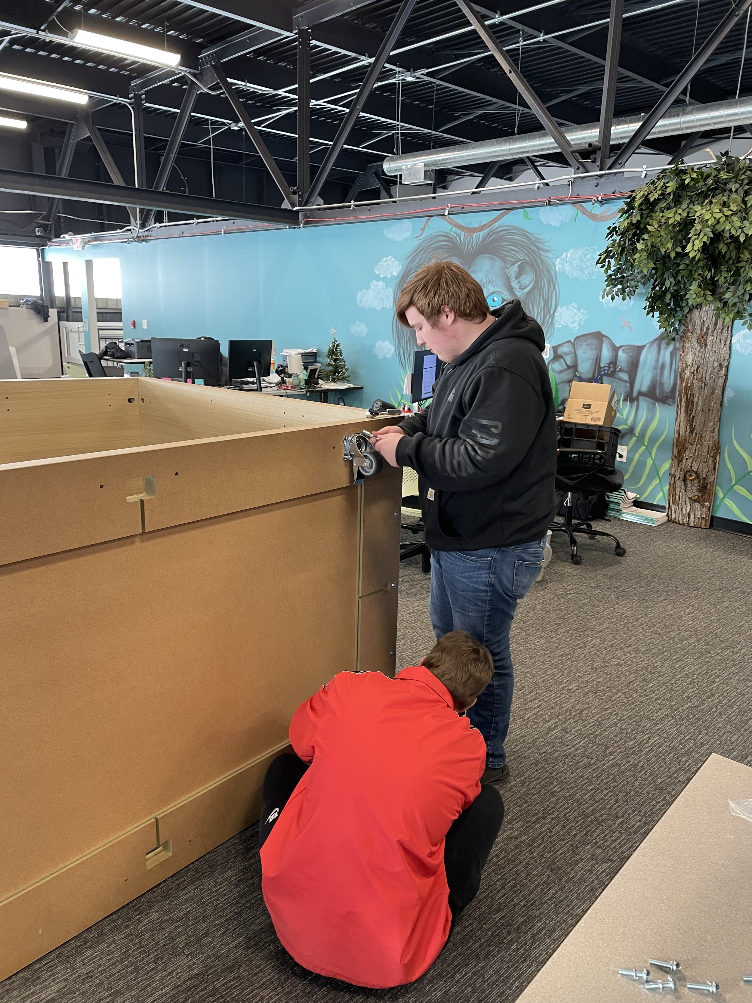 Two people assembling a large piece of furniture or equipment in an office setting with an artistic mural on the wall.