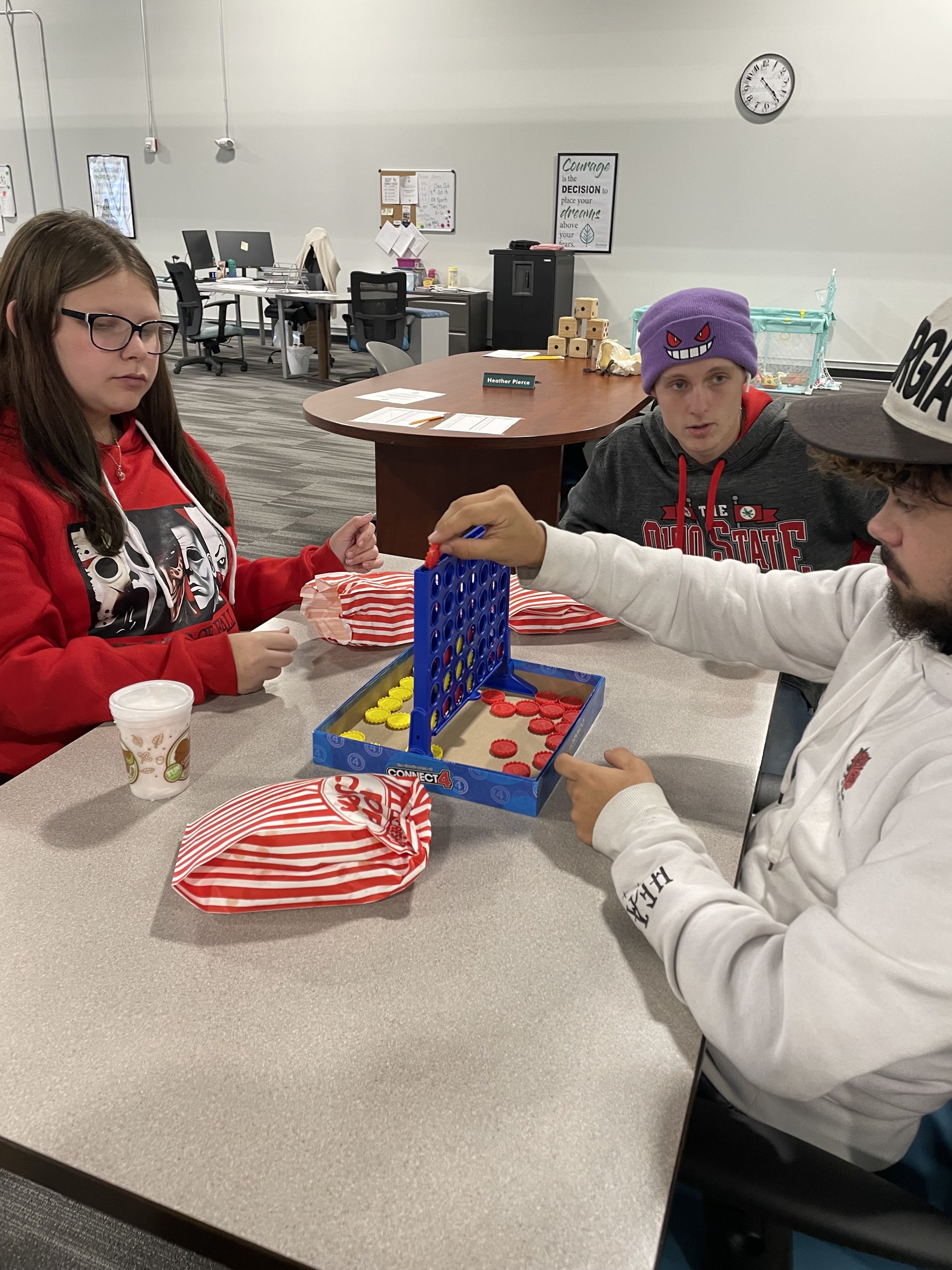Three people playing Connect 4 at a table in an office setting with snacks and drinks.