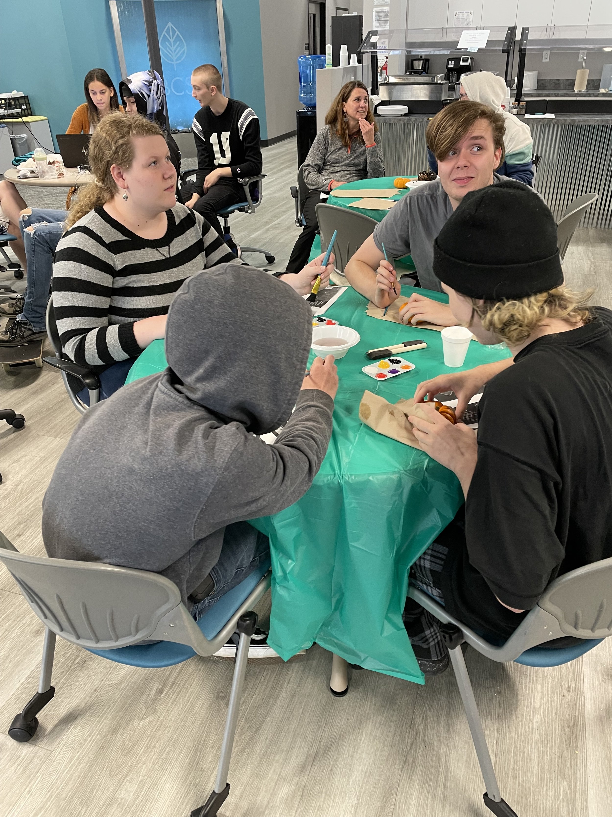 People sitting at tables engaged in a group activity or workshop in a casual setting, with some individuals painting on small canvases.