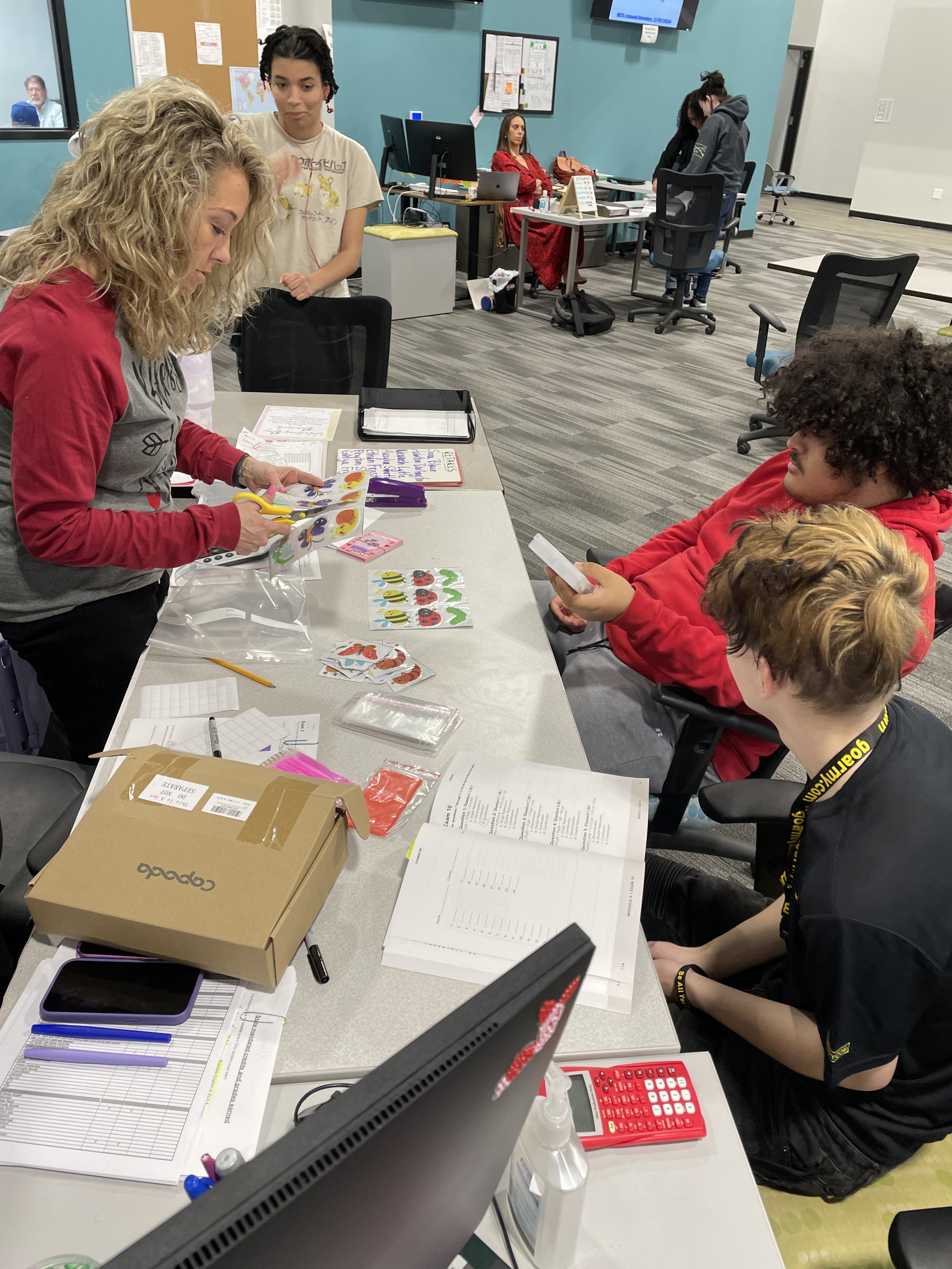 Students and a teacher in a classroom setting, working at a table with books, papers, and school supplies.