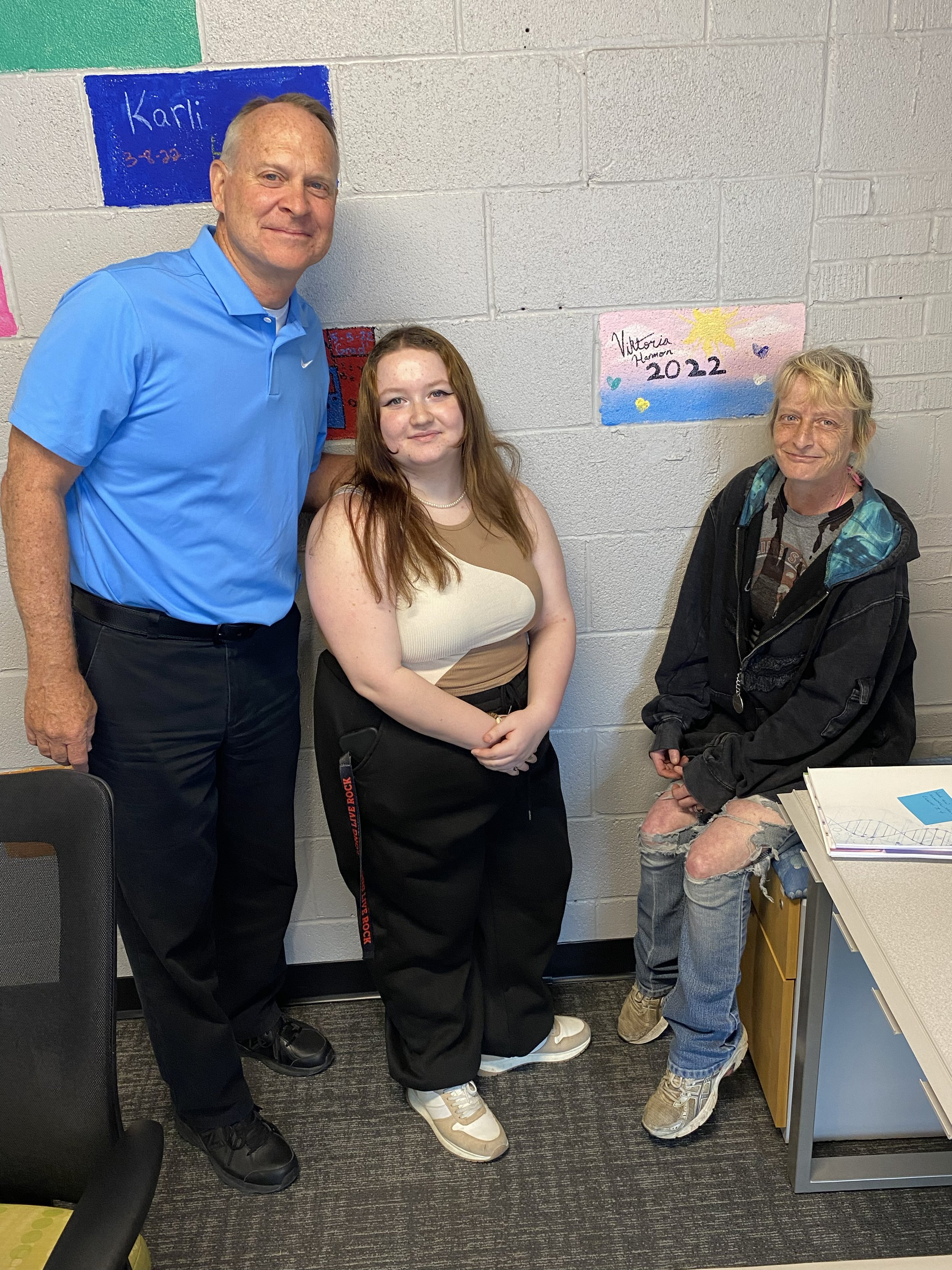 Three people posing together in a room with painted bricks on the wall. One person is standing, another is leaning against the wall, and the third is seated on a cabinet. The setting appears casual and informal.