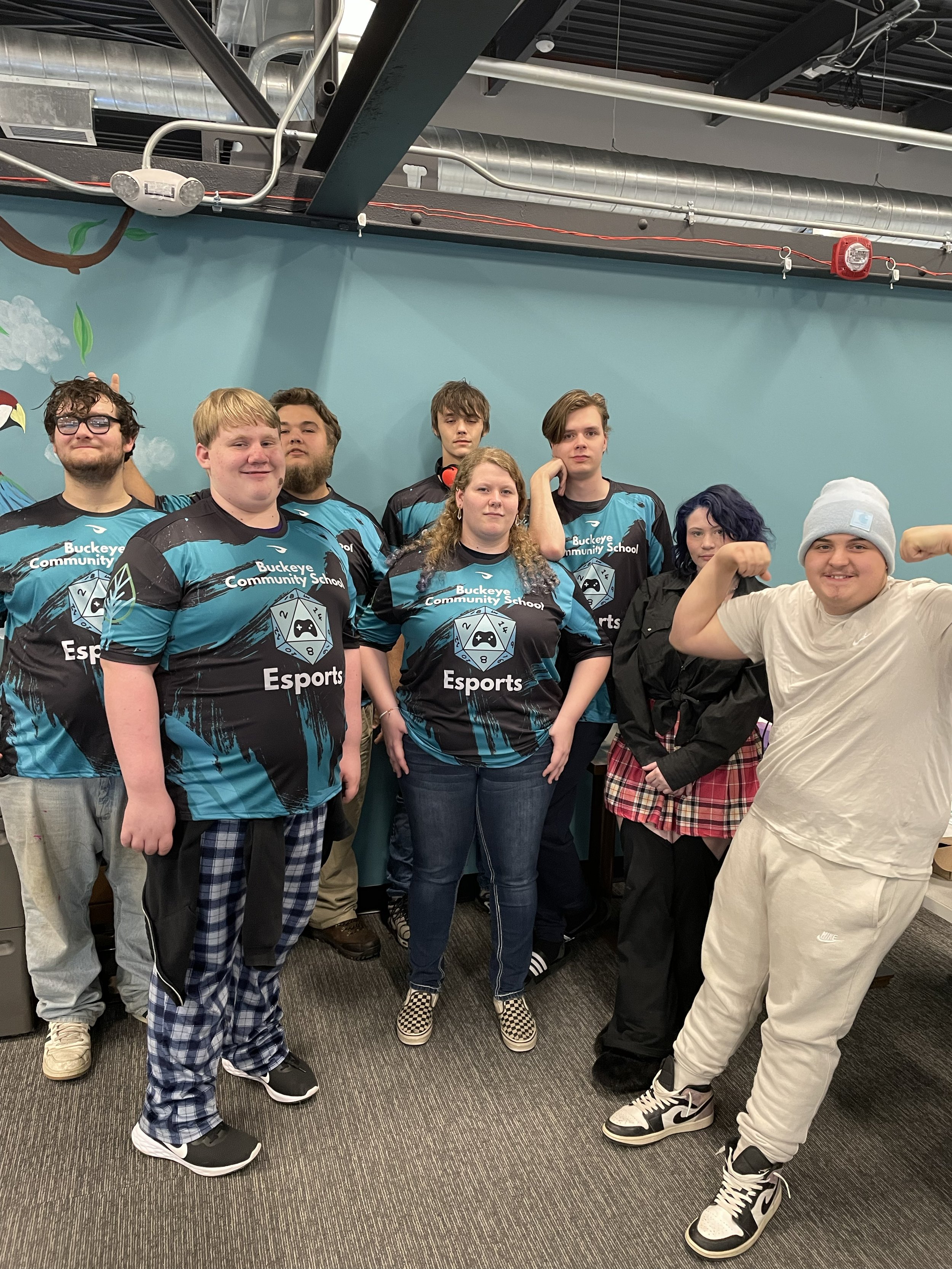 Group of students in esports team jerseys posing for a photo in a classroom setting with a teal wall in the background.