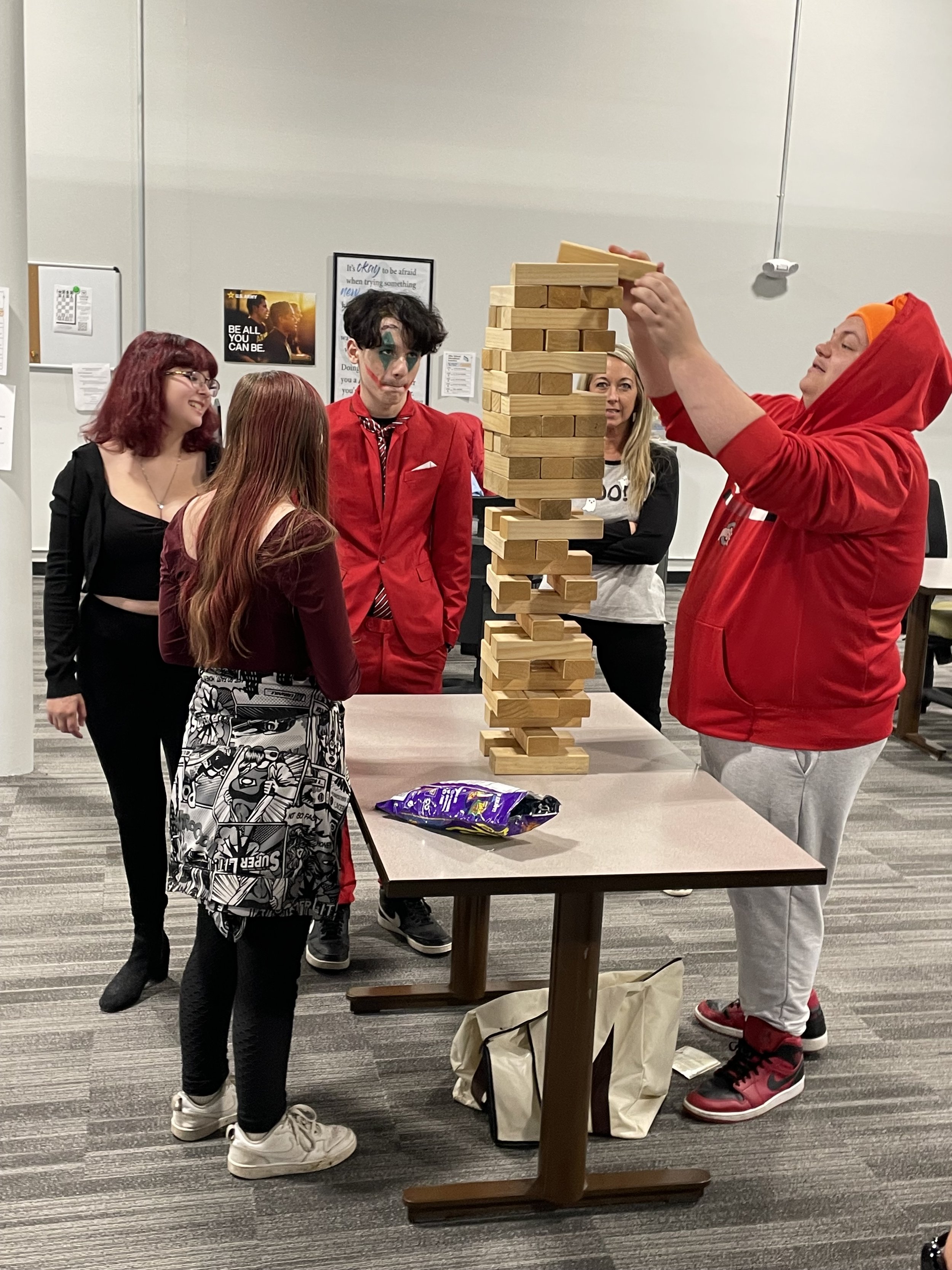 Group of young people playing Jenga on a table. One person is adding a block to the tower while others watch. They are in a room with posters on the walls and a snack bag on the table.