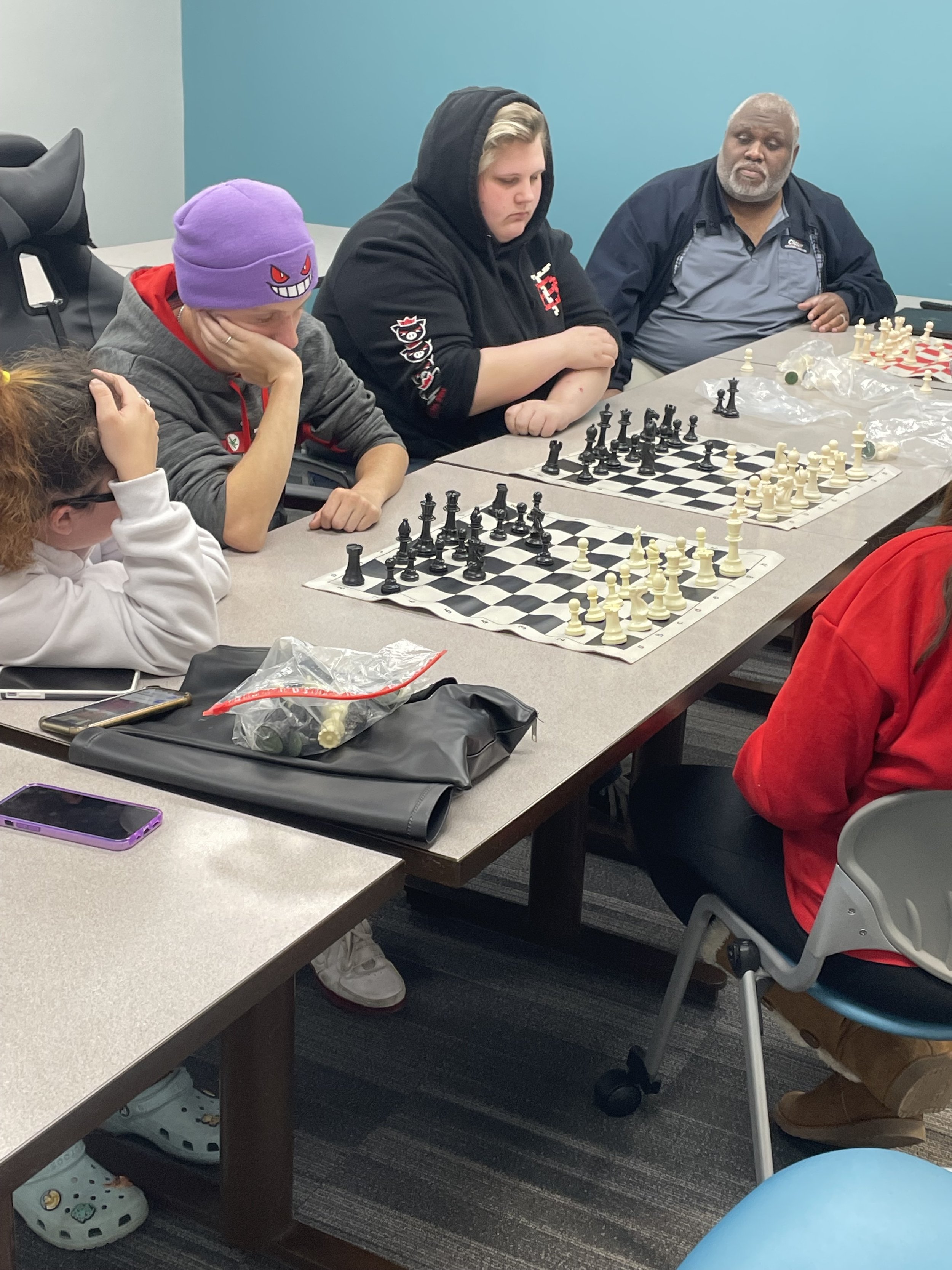 People playing chess in a room, focused on their games. Several chess boards are set on the table, and the participants are engaged in thought. A turquoise wall is in the background.