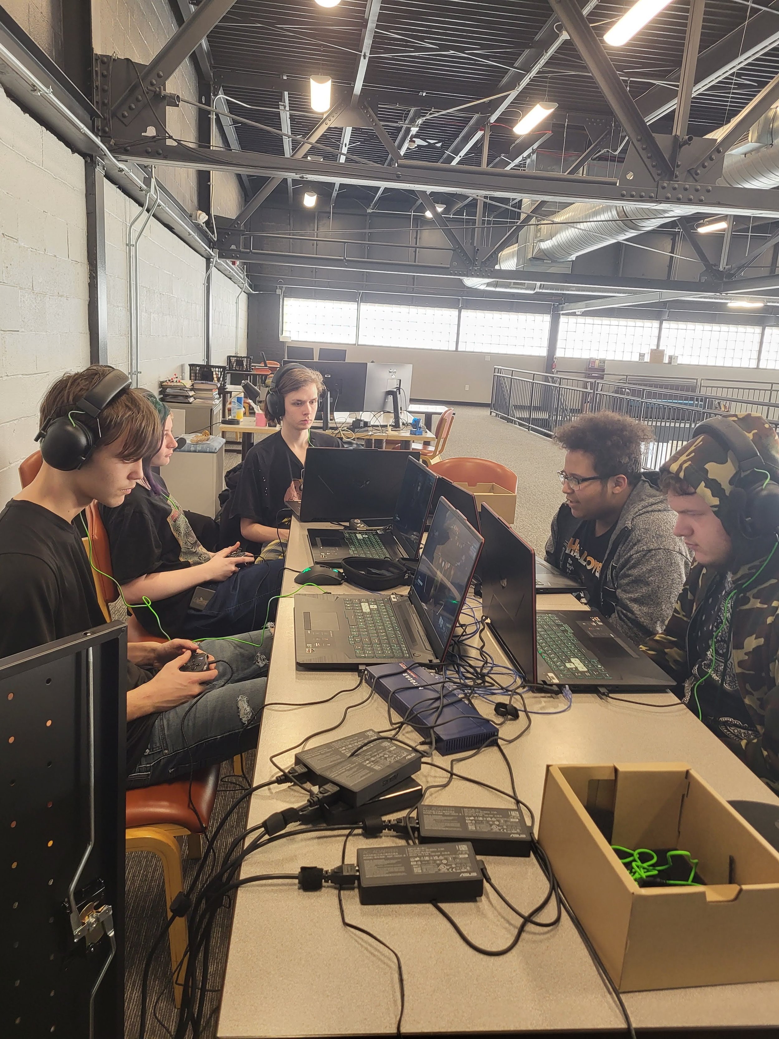Group of people gaming at a table with laptops and consoles in an industrial-style room, wearing headphones.