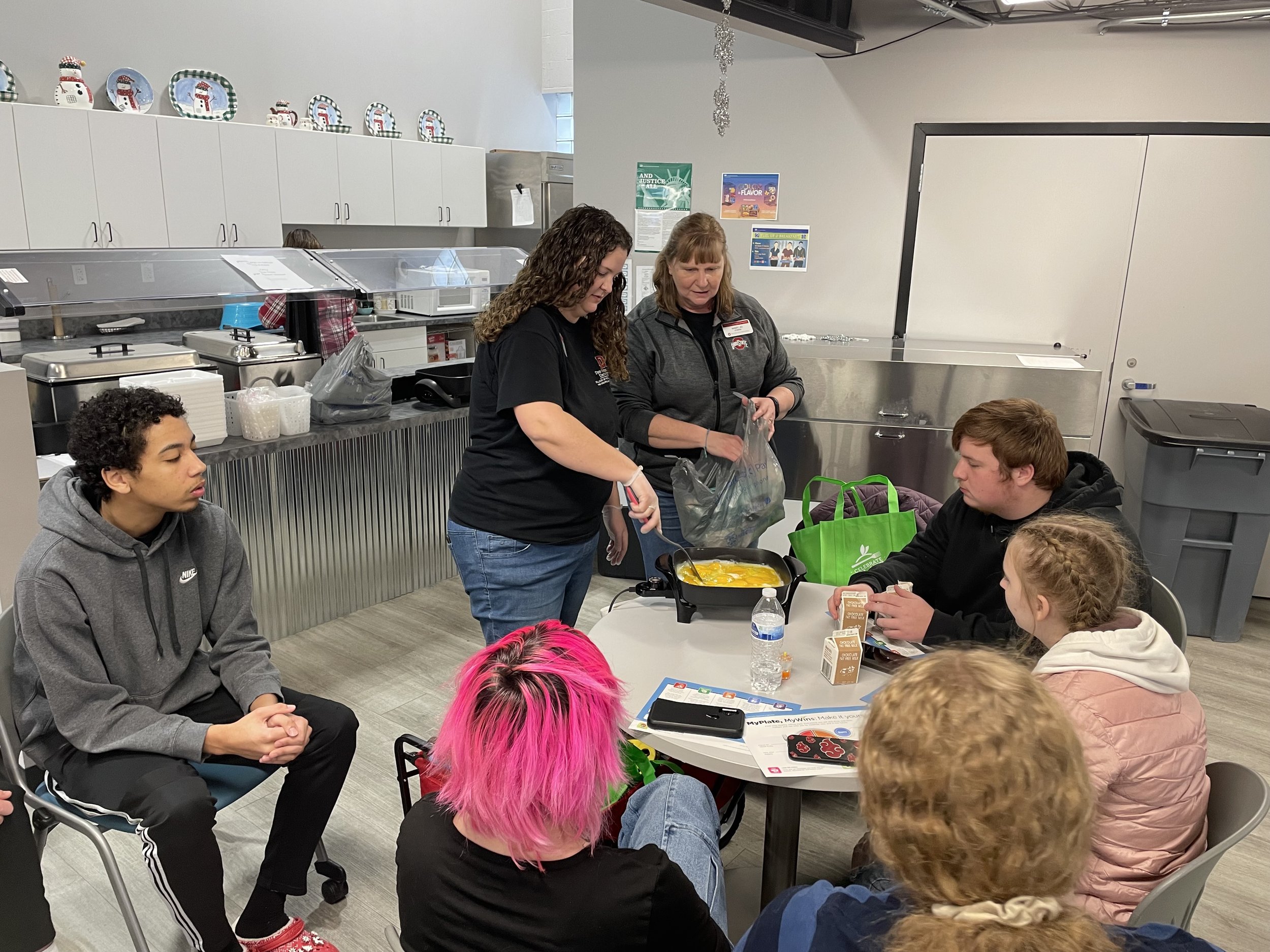 A group of people gathered around a table in a kitchen setting, with two women demonstrating cooking techniques using a frying pan. Several people are seated, watching and listening intently. The kitchen has white cabinets, stainless steel appliances