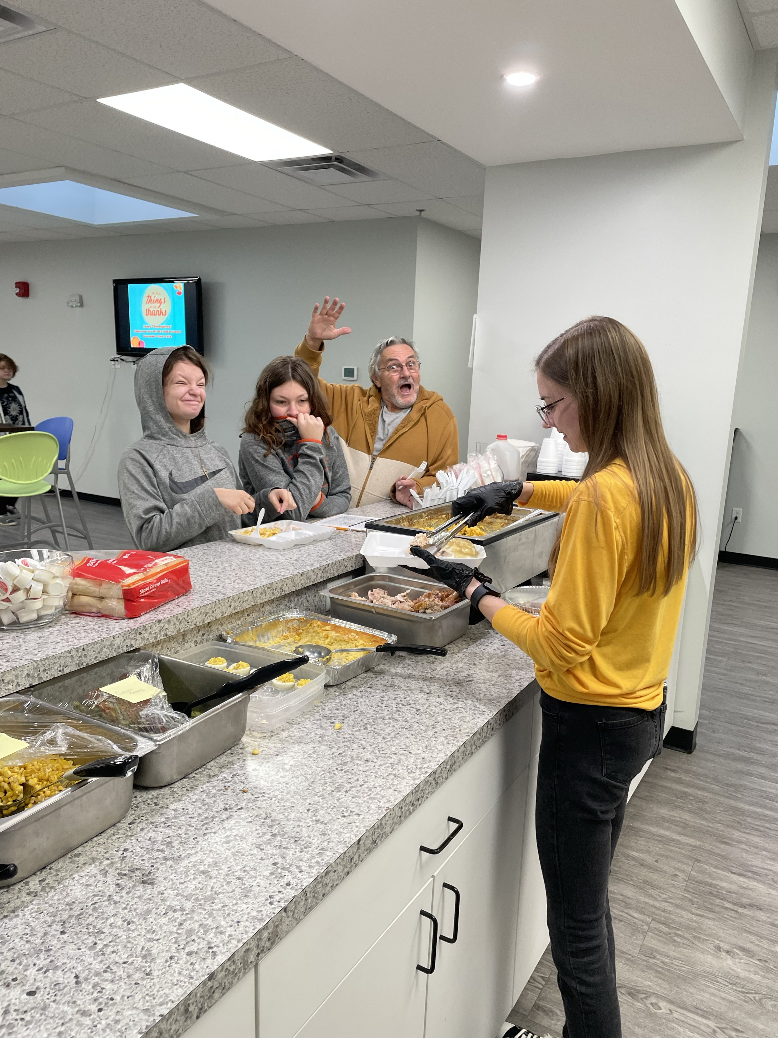 A group of people at a cafeteria counter serving and receiving food. A person with a yellow top serves food from trays filled with various dishes, while others stand on the opposite side of the counter, some smiling and one waving towards the camera.