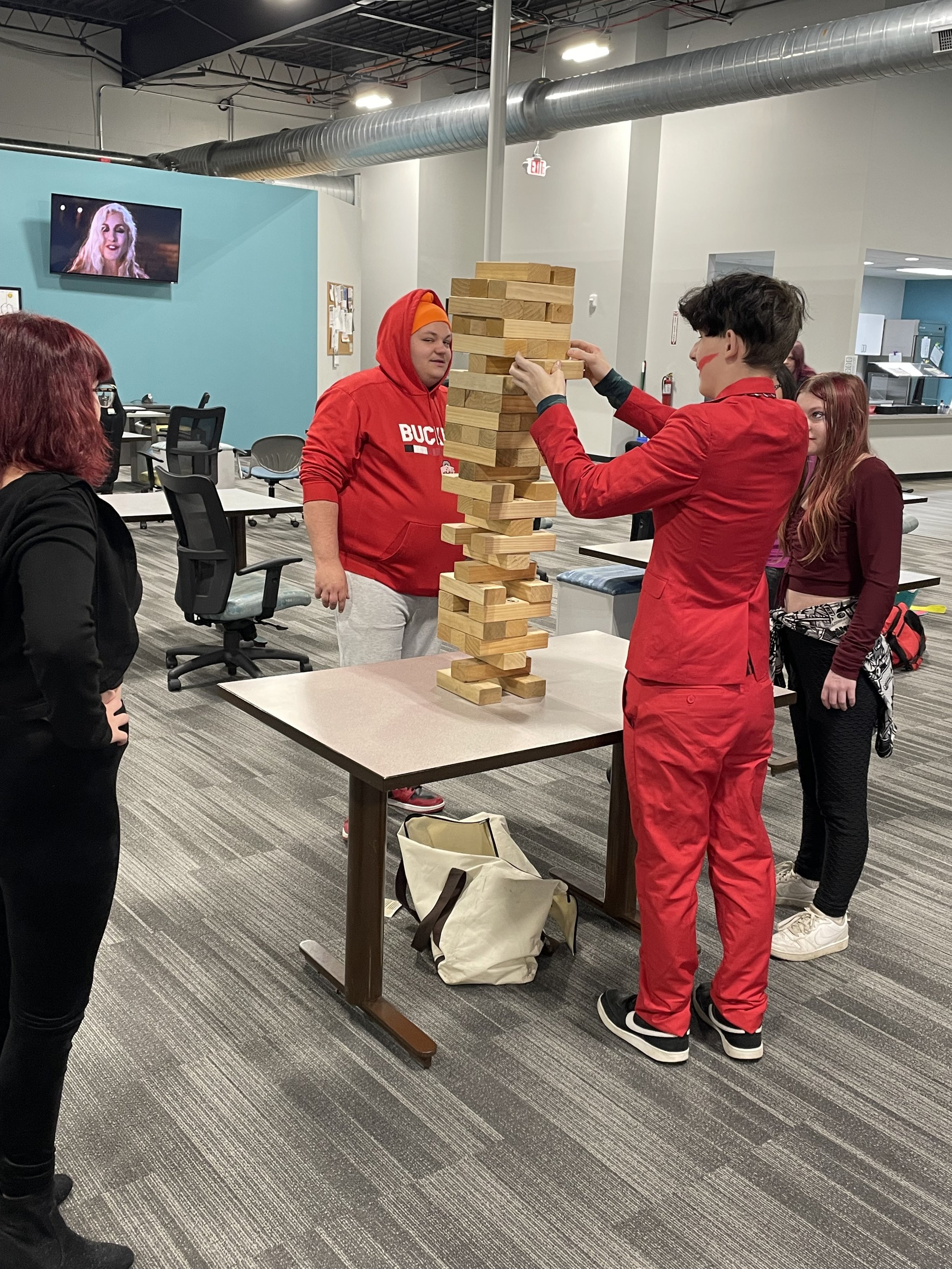 People playing a giant Jenga game in a modern office space with a TV on the wall in the background.