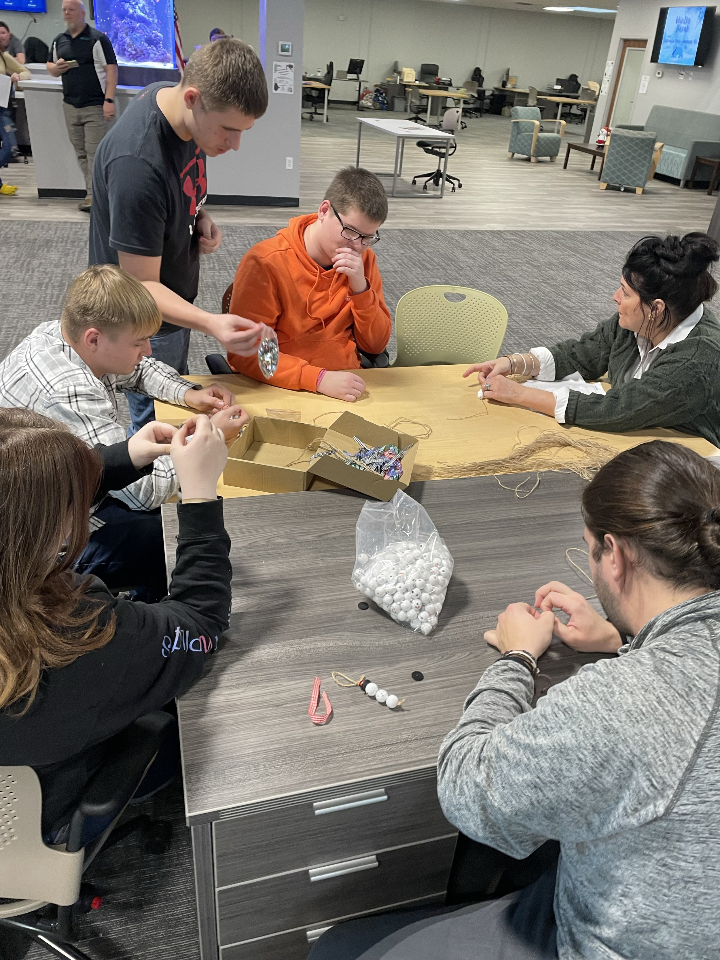A group of people sitting and standing around a table, engaged in a craft activity. There are various supplies on the table, including beads, string, and other crafting materials. The room is a spacious indoor area with office furniture in the backgr