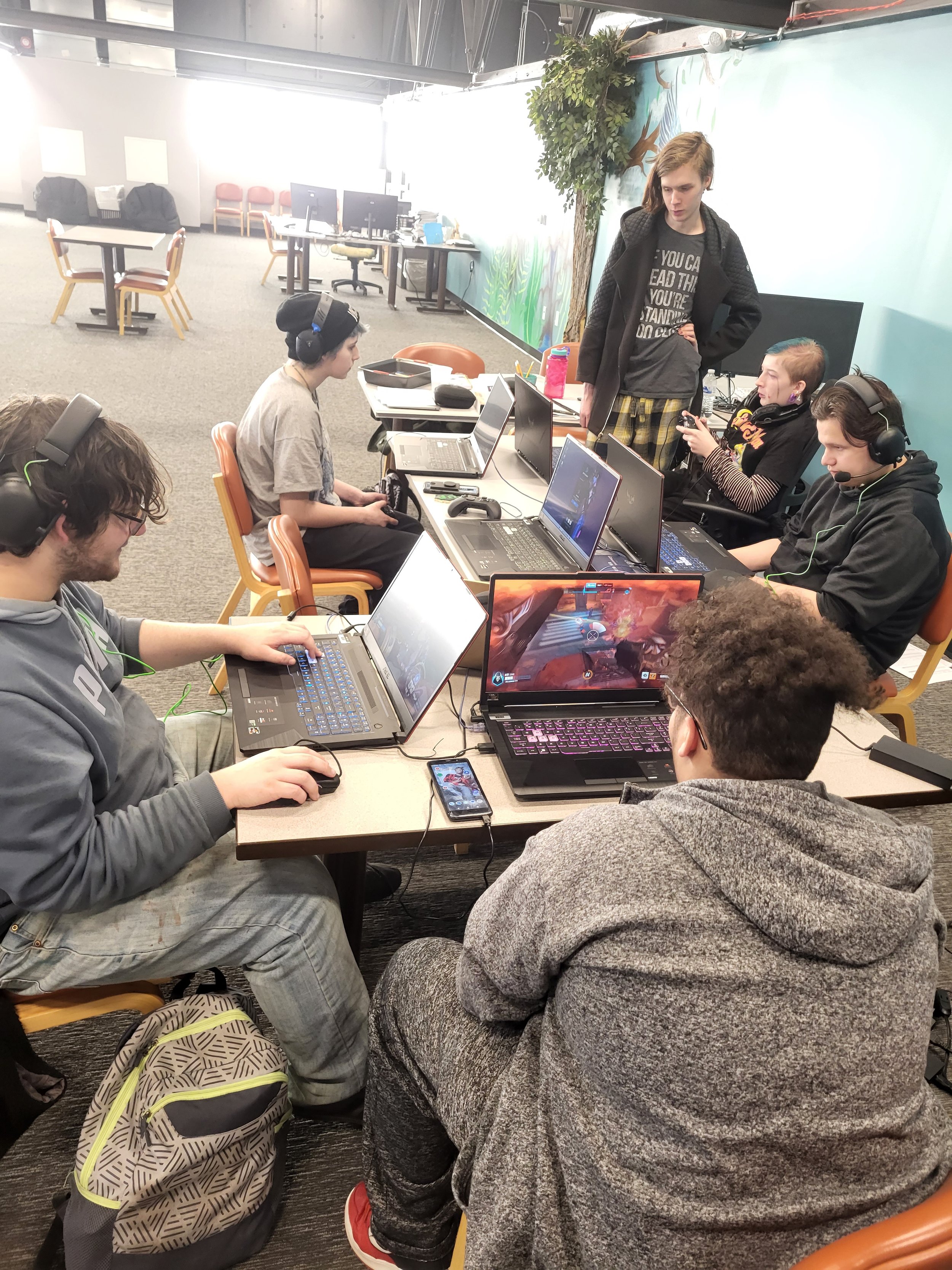 A group of people sitting at a table, engaged in video gaming on laptops. They are wearing headphones and using various gaming accessories. The room has a casual setting with tables, chairs, and computer equipment.