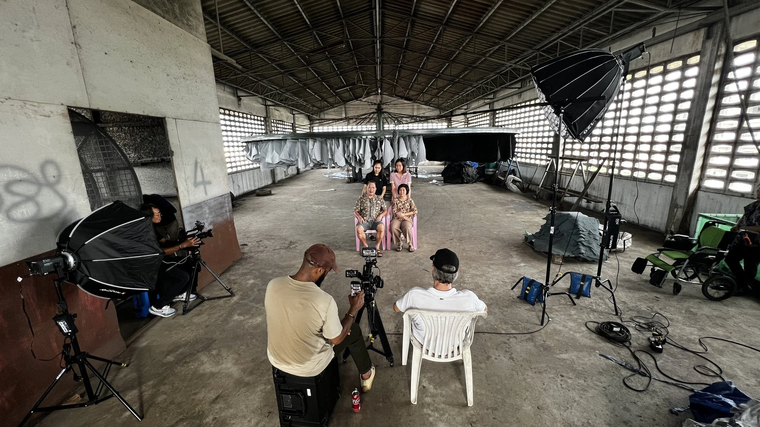 People filming a video or photoshoot inside a large, unfinished, open-air structure with a high, lattice roof, using professional studio lighting and cameras, with two individuals seated in pink chairs as models or interviewees and others standing or sitting around the scene.