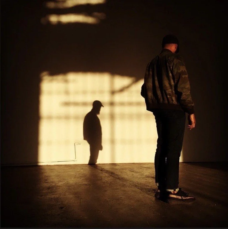 A person standing in front of a wall with their shadow projected on it, showing a person with a cap. The light creates an elongated shadow of the individual on the wall.