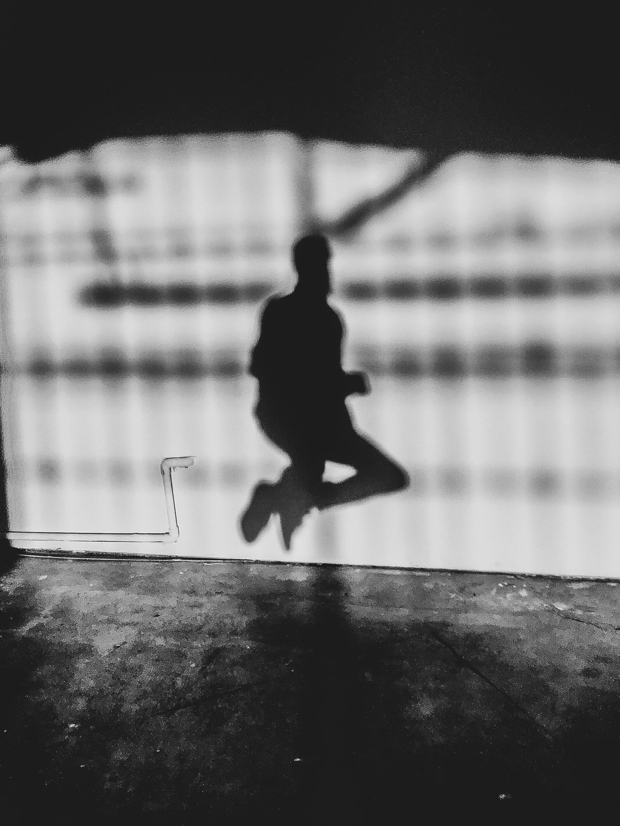 Silhouette of a person sitting on a suspended swing or ledge, with a blurred background of what appears to be a building or industrial structure, in black and white.