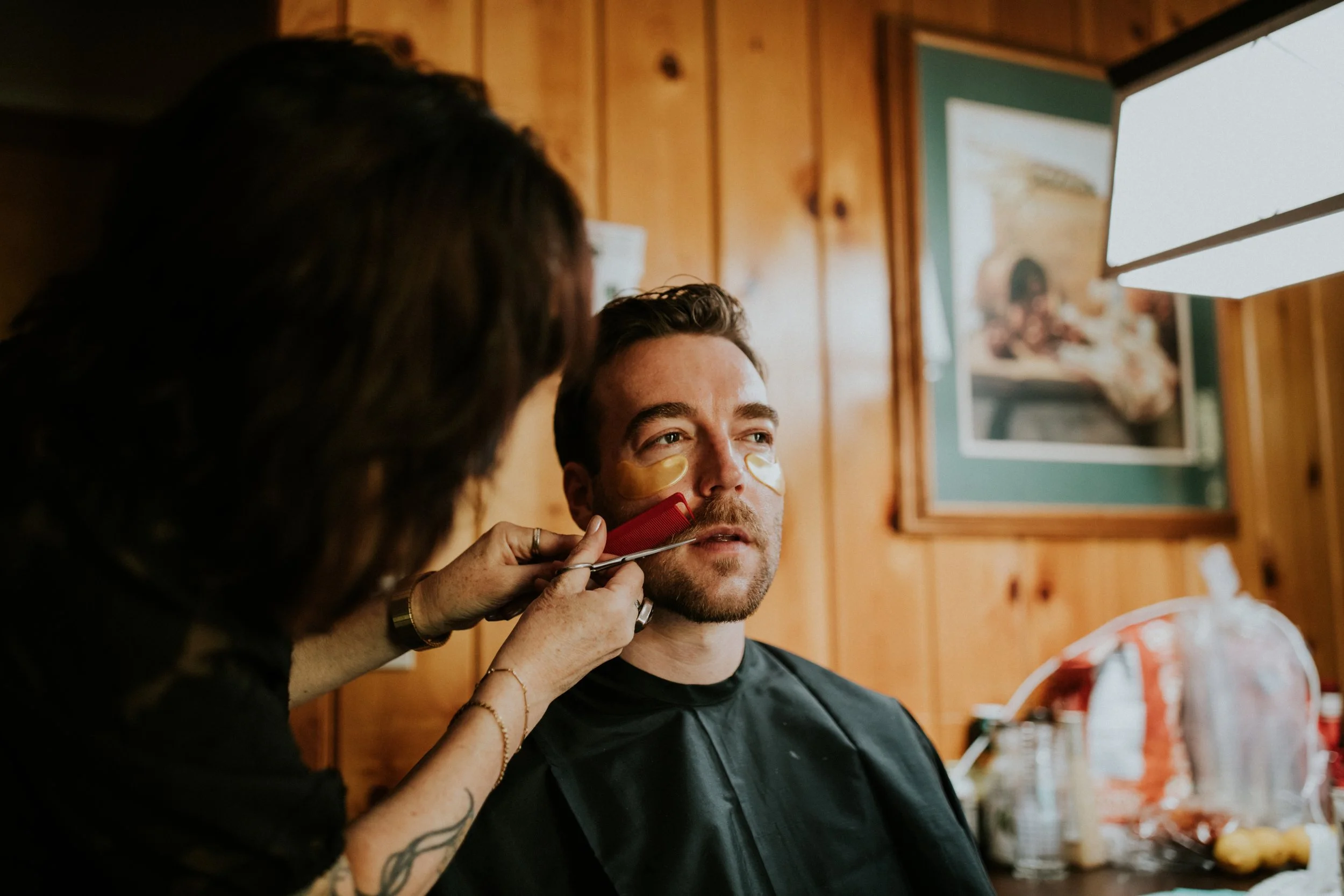 A gay man getting a wedding day glow up by a woman in a rustic wooden room, with a framed picture on the wall by Dorey Kronick