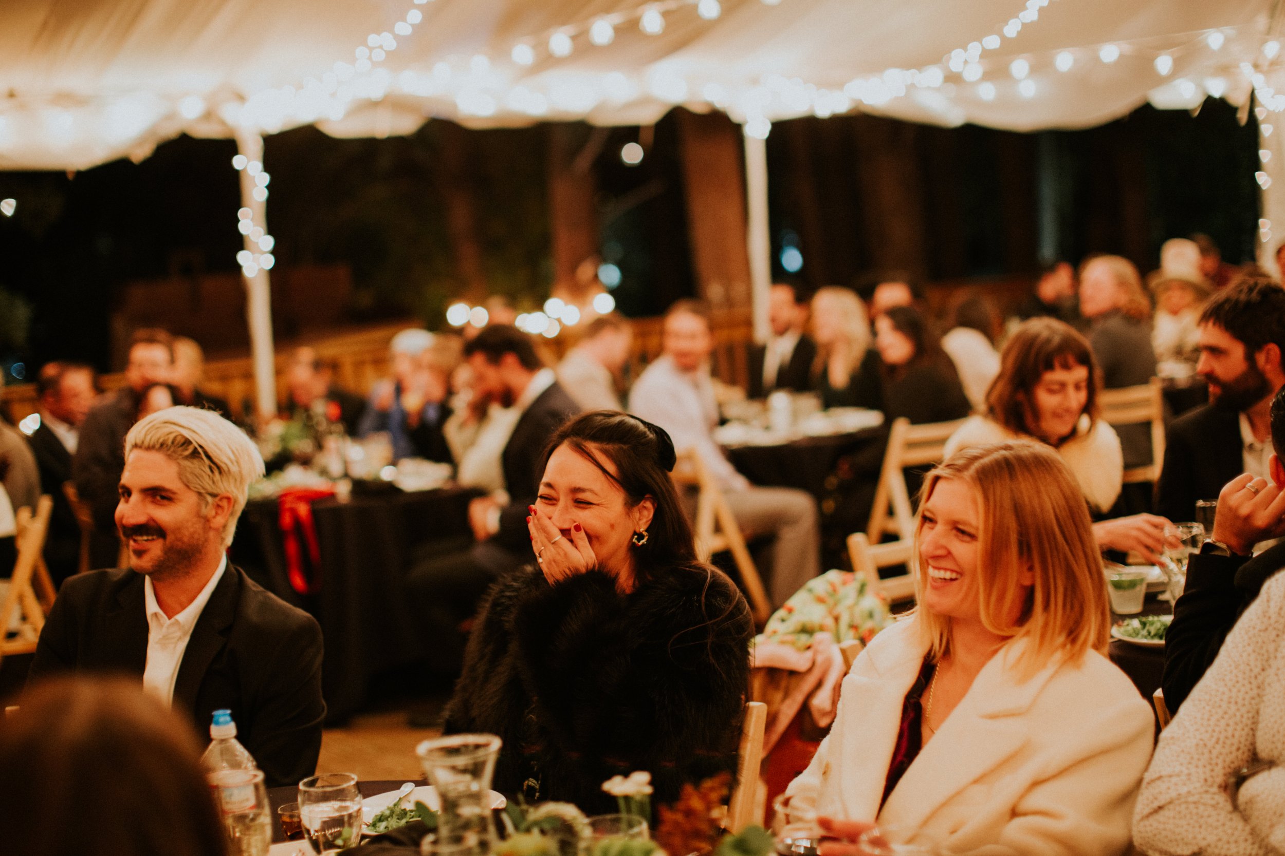 People seated at tables under string lights at an outdoor evening wedding reception in the fall, smiling and laughing by Dorey Kronick