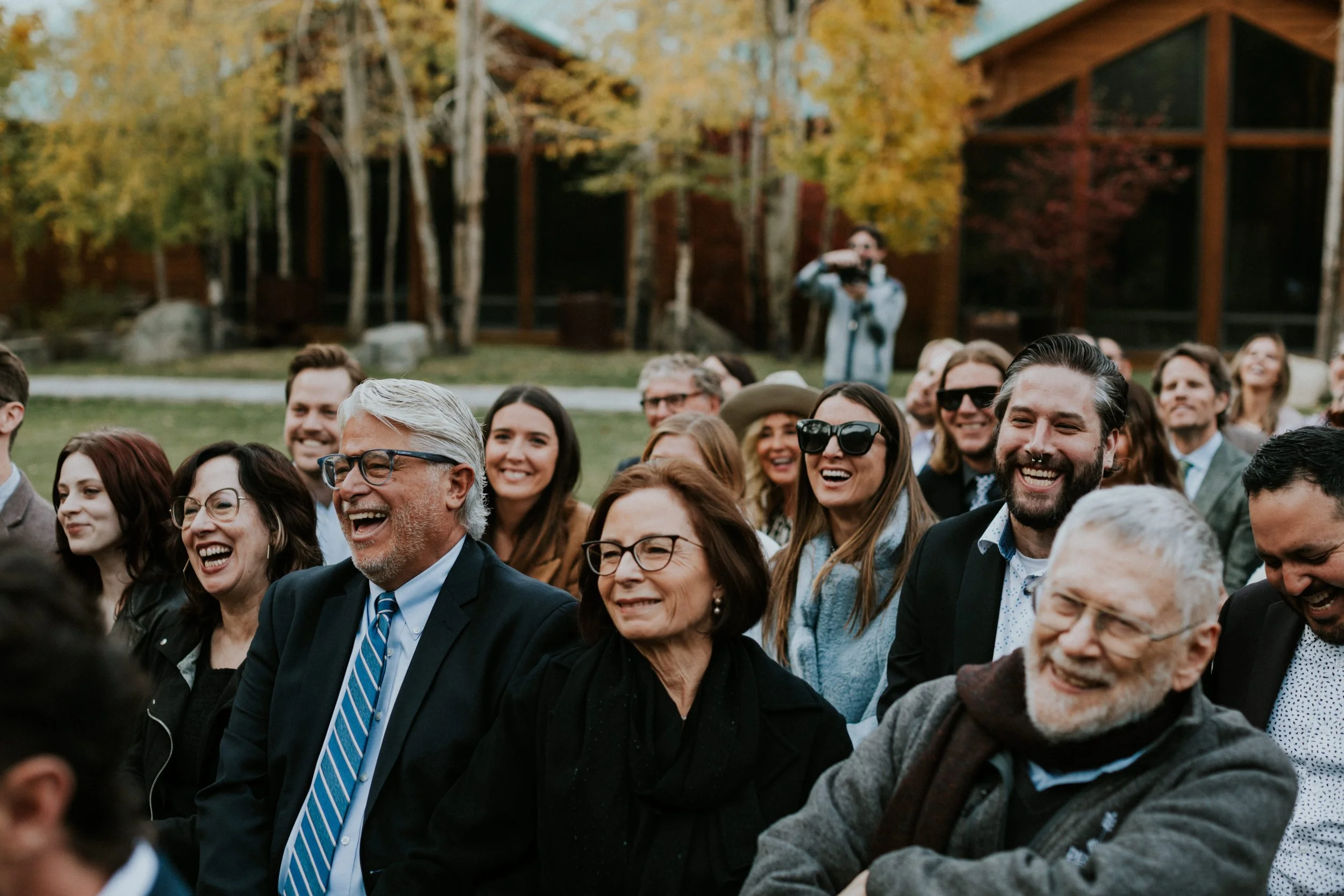 A group of people sitting outdoors, smiling and laughing during a gathering in a park with autumn-colored trees.