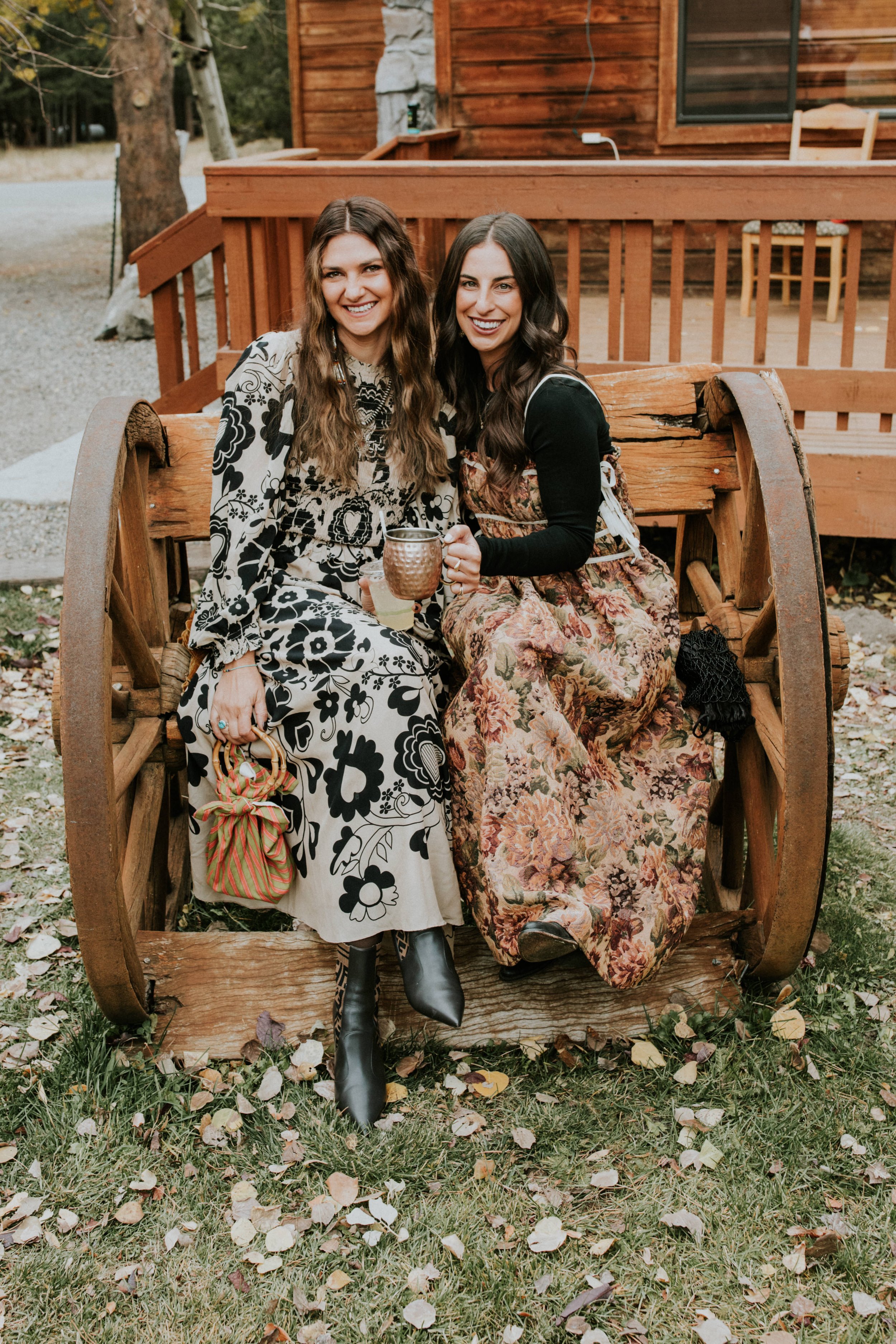 Two women sitting on a wooden cart, smiling and holding drinks, outdoors in a yard with fallen leaves and wooden cabin in Mammoth, CA by Dorey Kronick