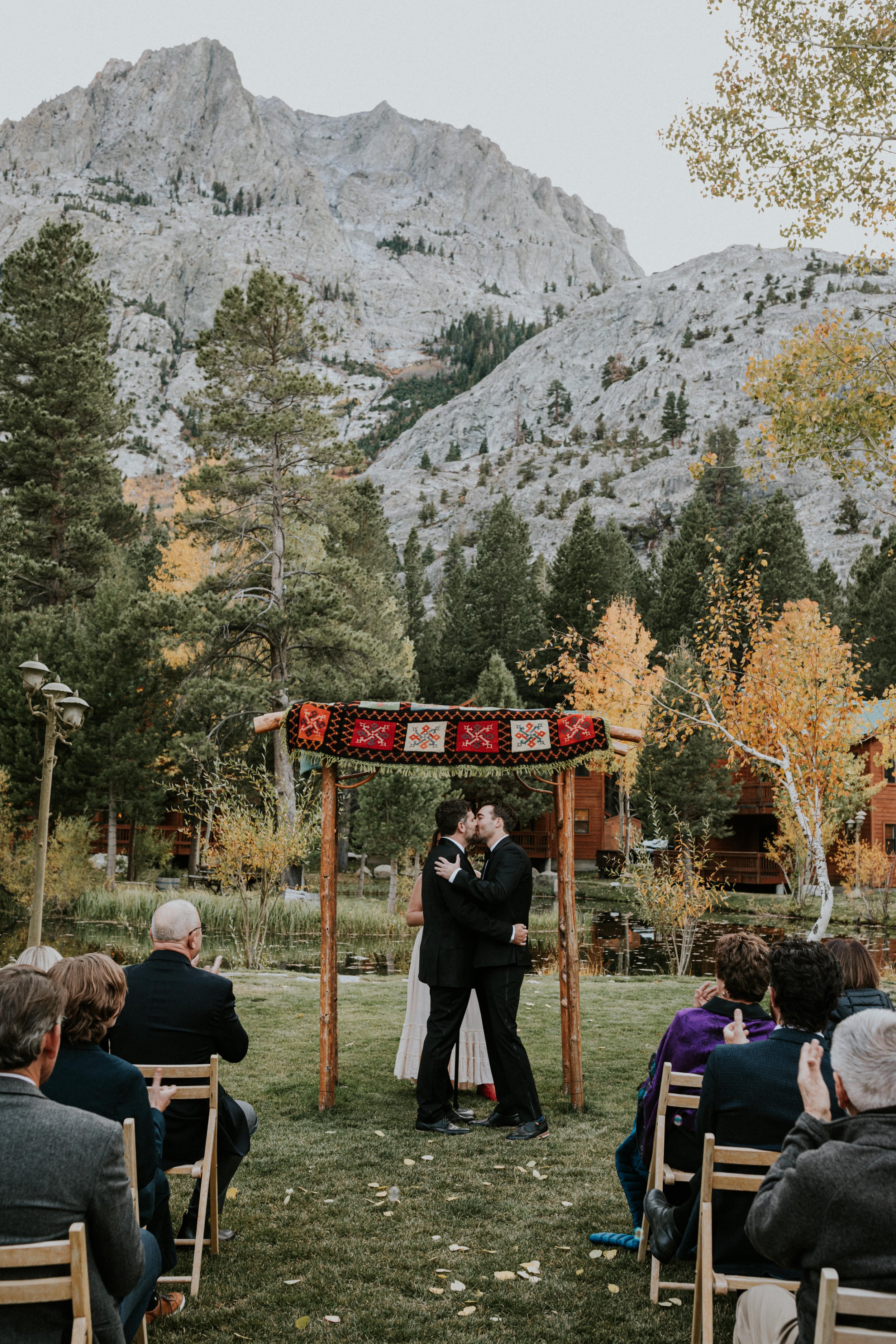 Two men in tuxedos kissing under a decorated canopy at an outdoor wedding ceremony with mountains, trees, and a pond in the background.