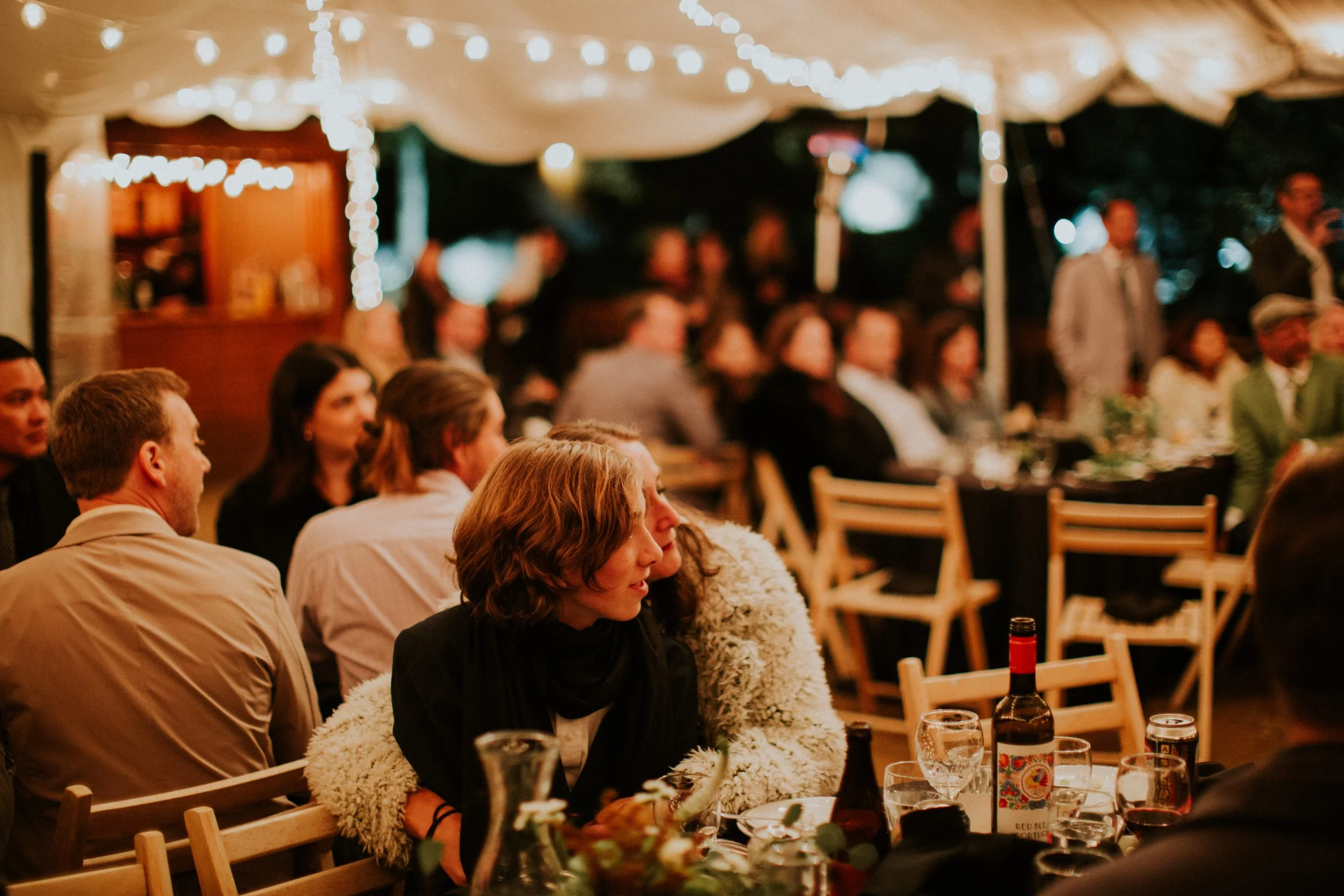 People sitting at tables under a decorated tent at night, attending an LGBQTIA+ wedding, with string lights overhead by Dorey Kronick