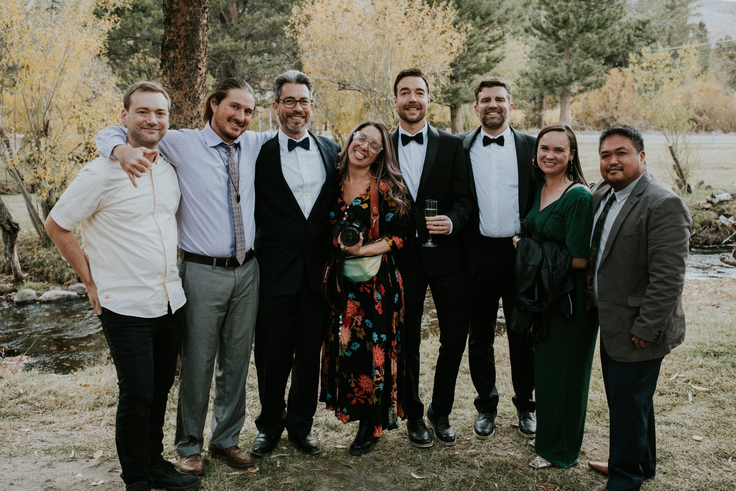 Group of nine dear friends outdoors, smiling and posing for a photo with well known designer, artist, and photographer, Dorey Kronick (holding camera), with some holding drinks, in California at a gay wedding