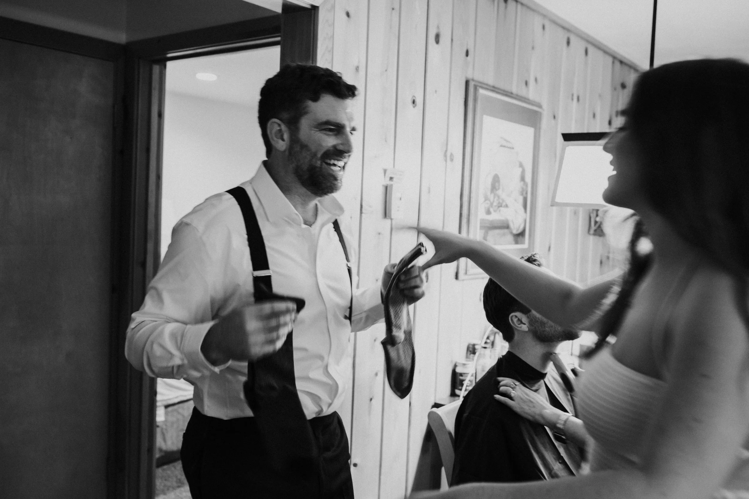 A gay man in a white shirt with suspenders holding a sock and a woman in a sleeveless top pointing at something in conversation inside a room with wooden panel walls by Dorey Kronick