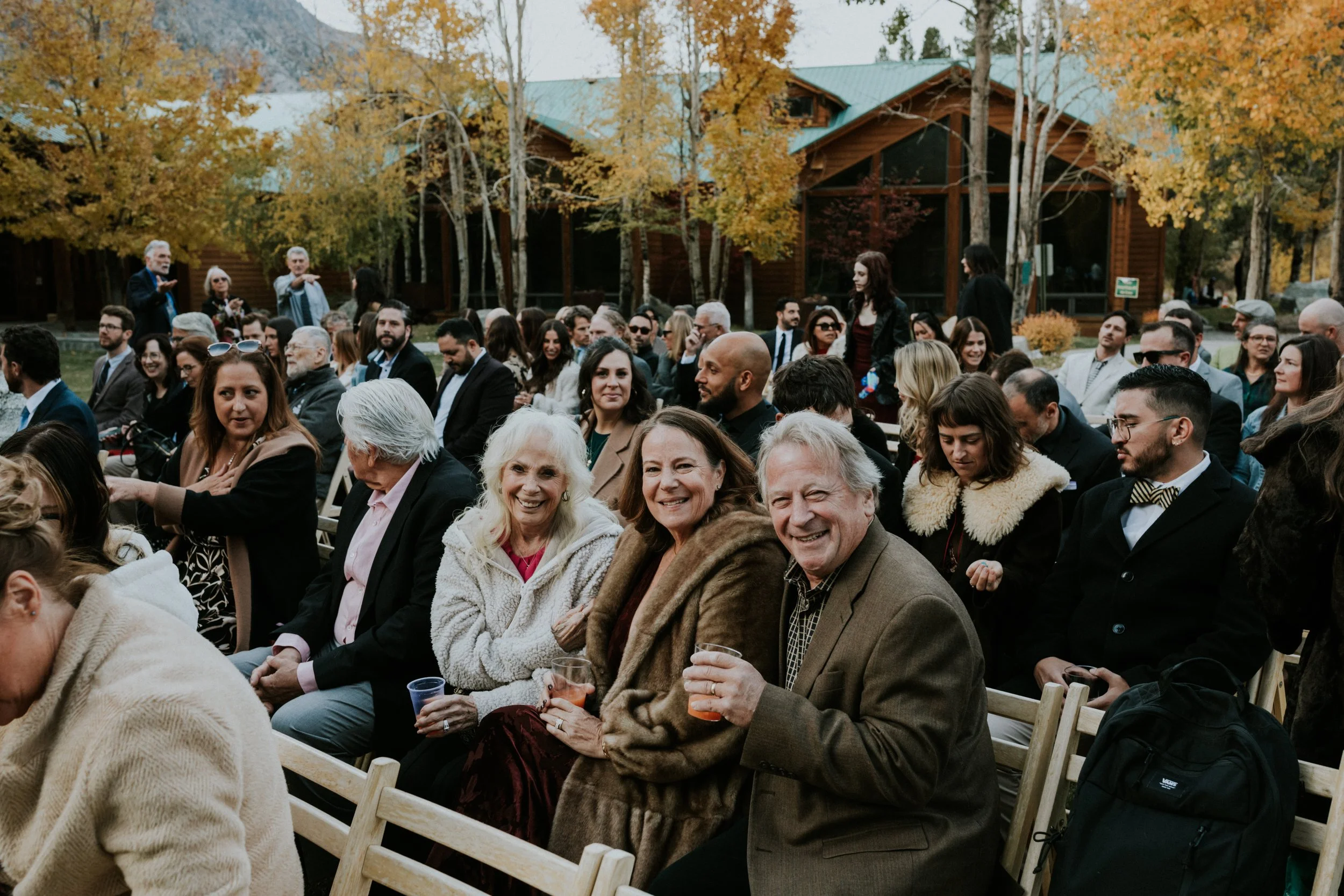 People sitting outdoors at a gay wedding, enjoying drinks, among autumn trees and wooden cabins in June Lake, CA by Dorey Kronick