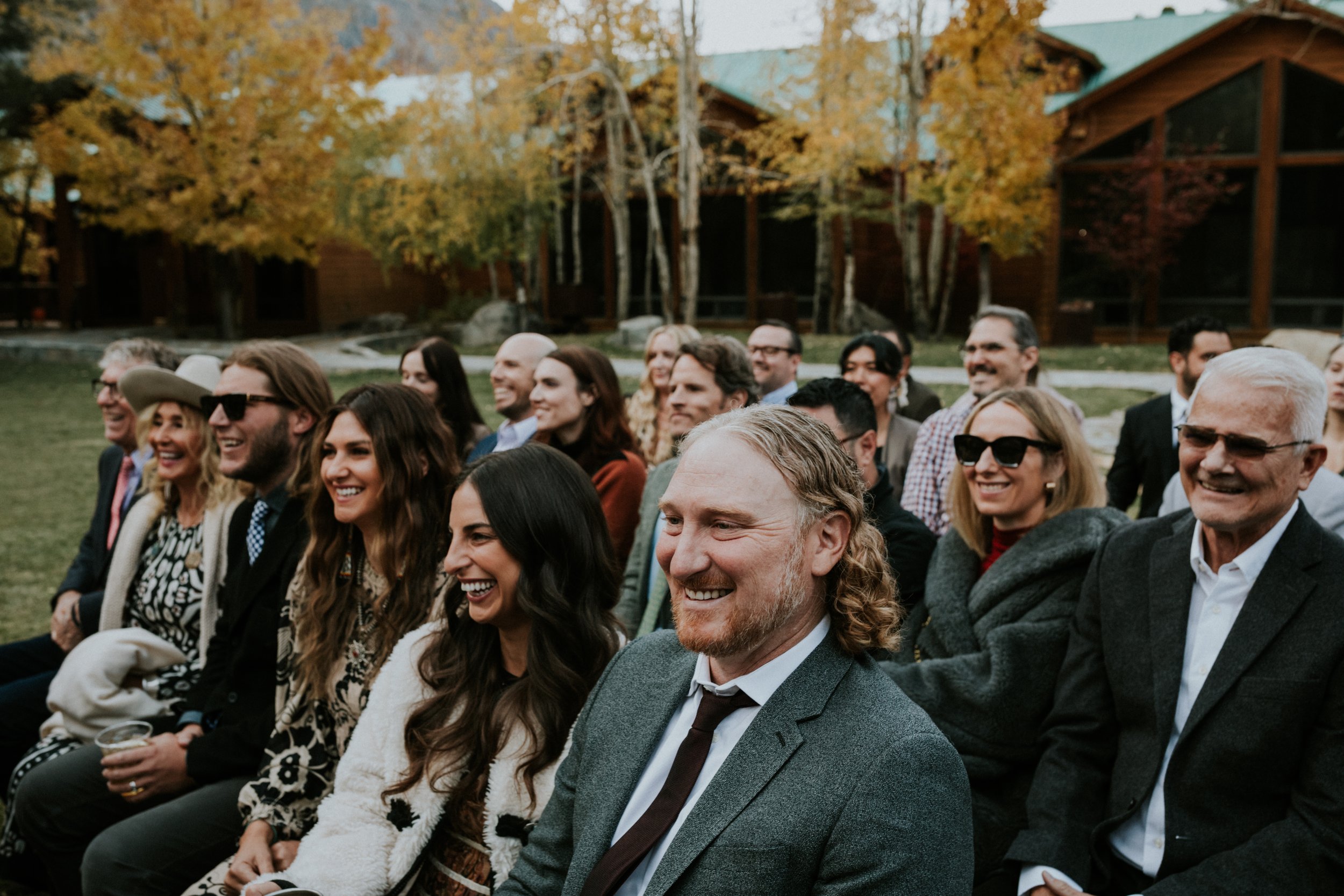 A group of people sitting outdoors, smiling and laughing during a social event, with autumn trees and a wooden building in the background at the Double Eagle Resort in June Lake, CA by Dorey Kronick