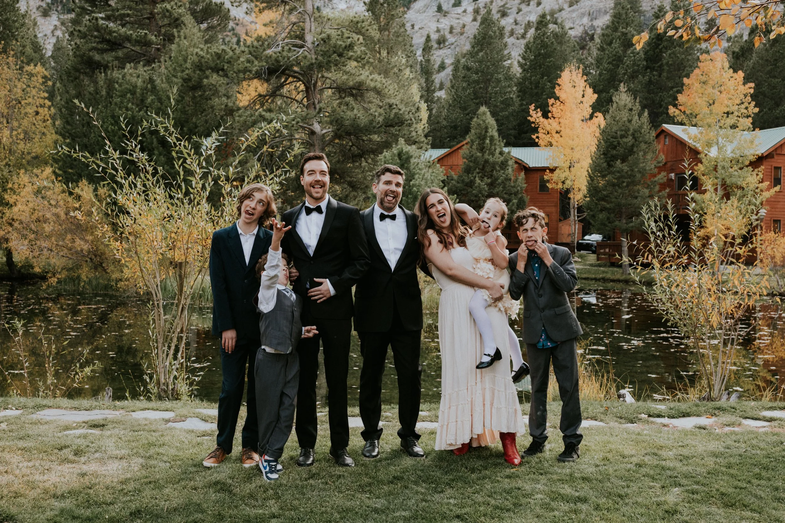 A group of seven people dressed in formal attire, standing by a pond with autumn-colored trees and wooden cabins in the background, celebrating at an outdoor wedding in June Lake, CA by Dorey Kronick
