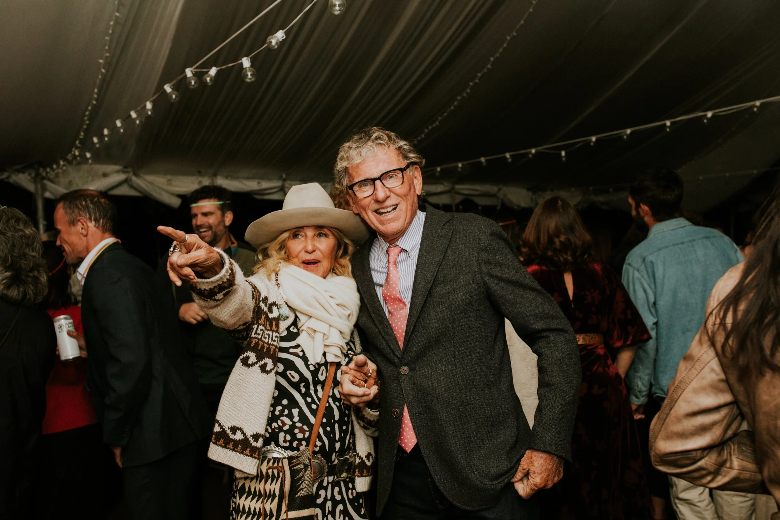 Two older adults, a woman wearing a large hat, stylish dress, and a scarf, and a man in a suit, smiling and holding hands at a crowded gay wedding under a tent with string lights at a resort in California by Dorey Kronick