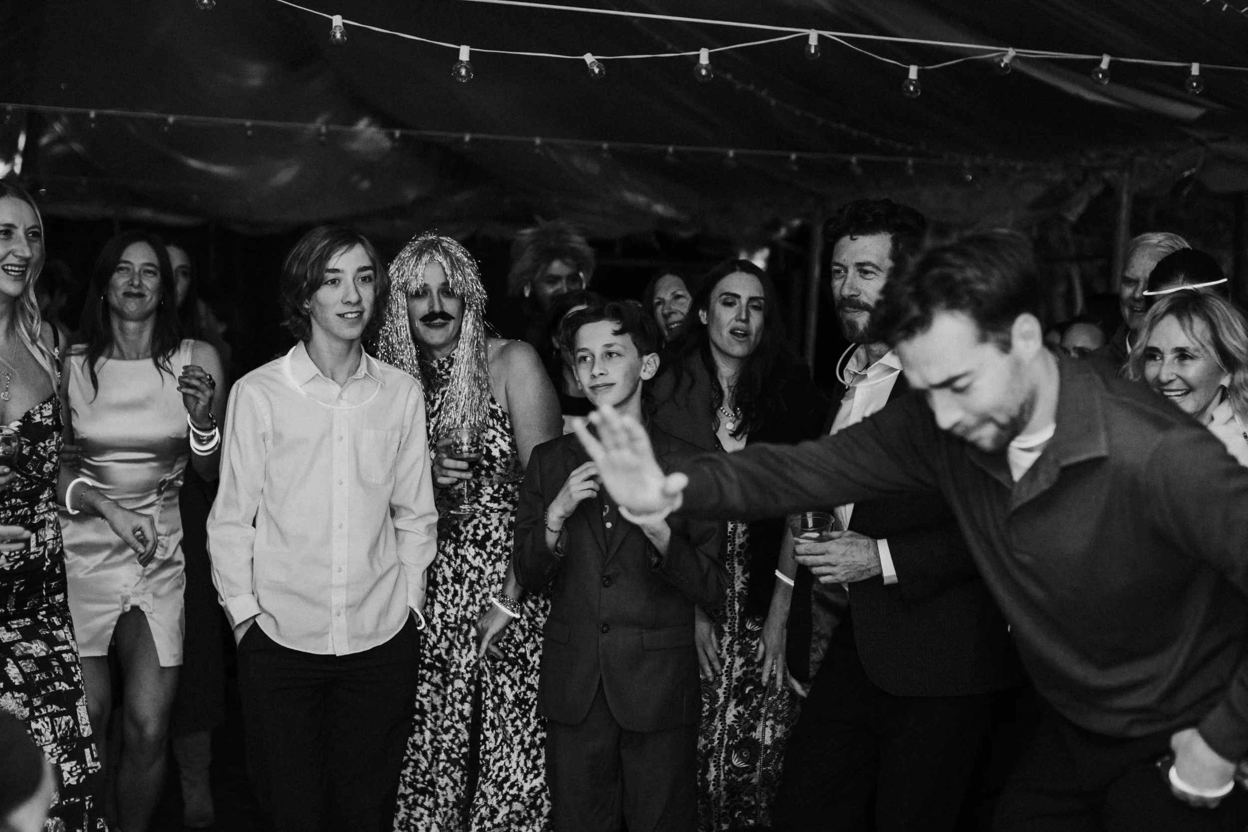 People dancing and socializing at a gay wedding reception under string lights, with one of the grooms in the foreground appearing to dance or perform by Dorey Kronick